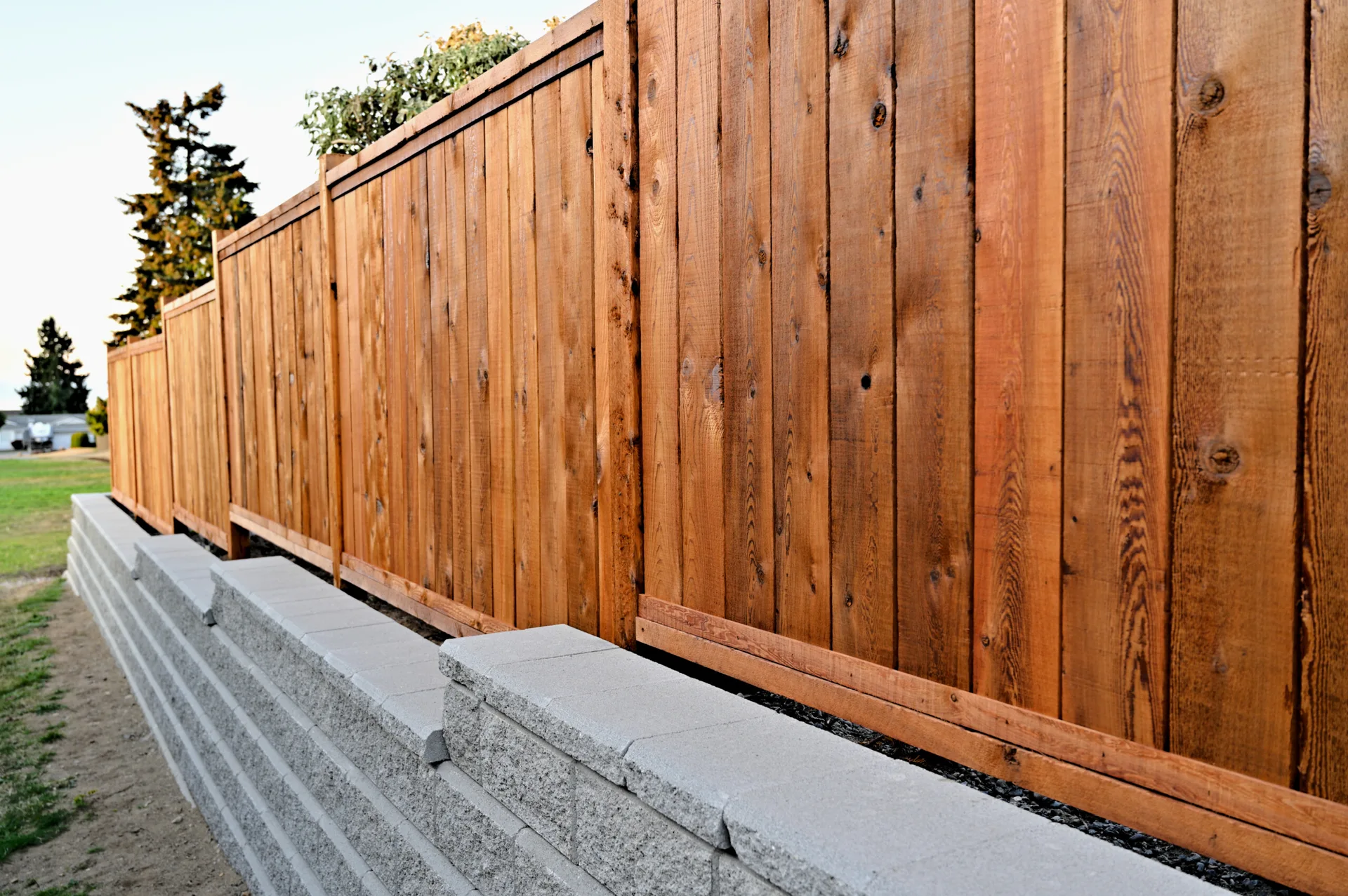 A tall stained wooden privacy fence stands behind a stepped grey concrete block retaining wall in a backyard landscape.