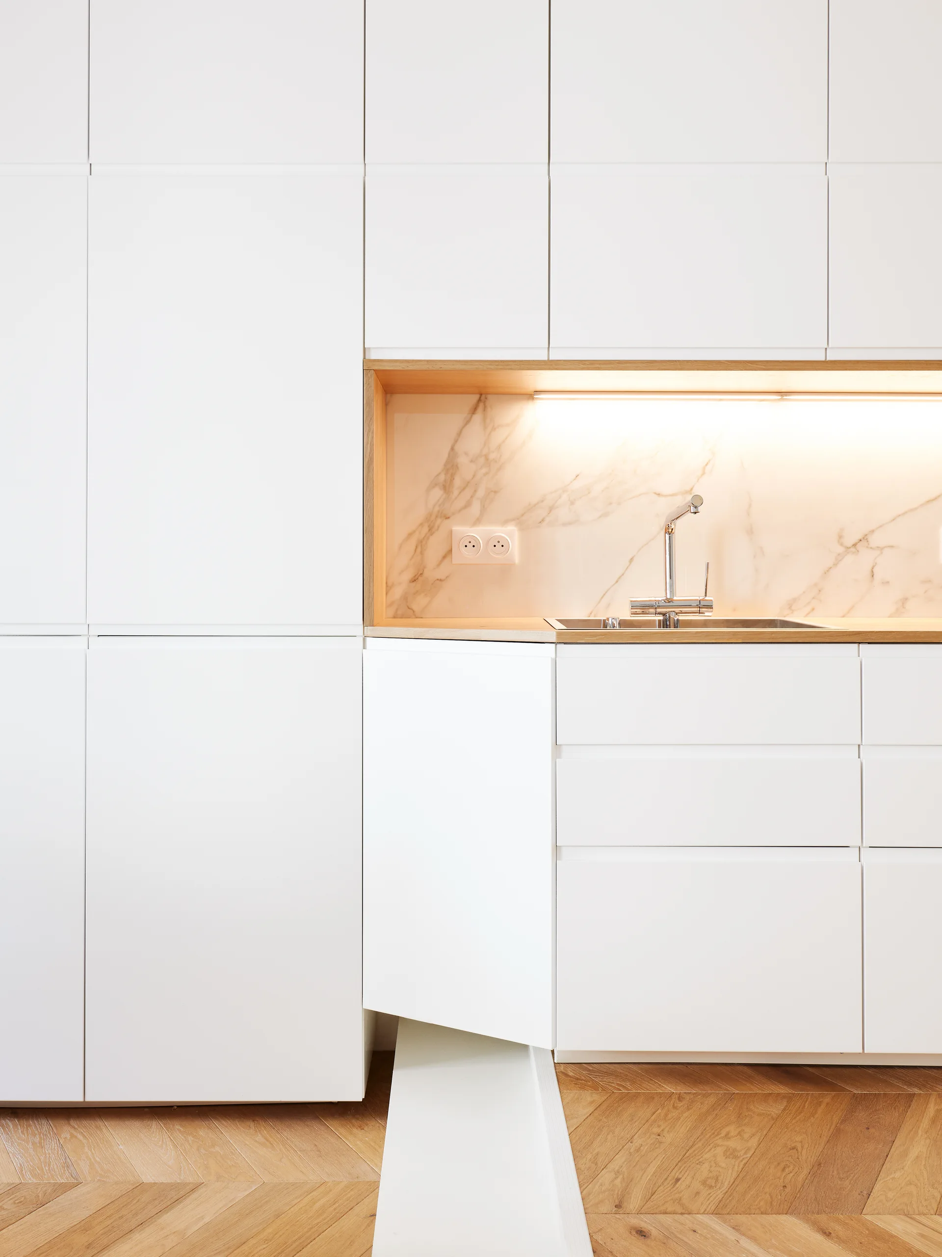 Minimalist white kitchen cabinets feature a recessed wood-lined niche with a marble backsplash, stainless steel sink, and light oak herringbone flooring.