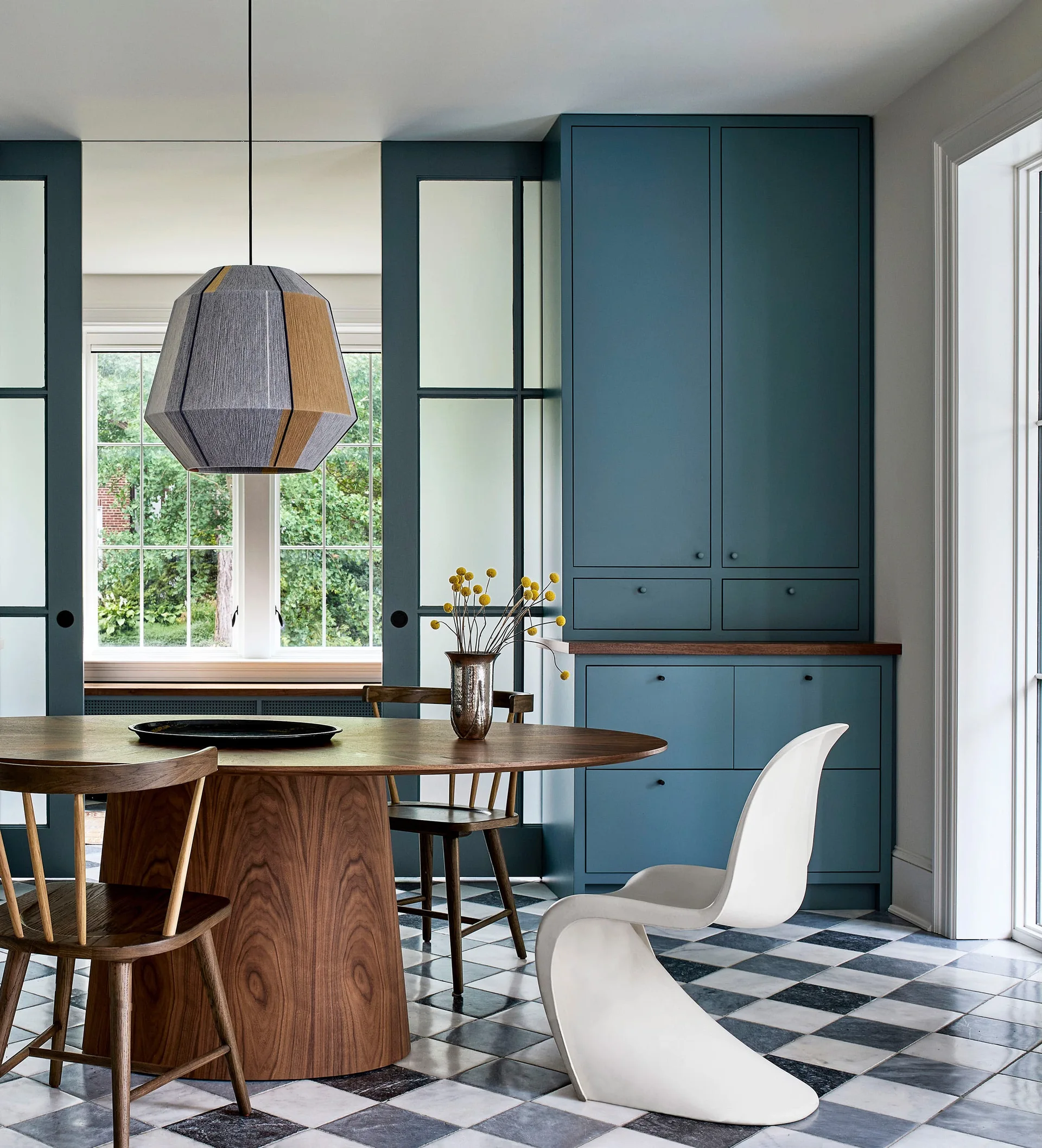 A round wooden dining table and mixed chairs set on a black and white checkered floor in front of custom blue built-in cabinetry.