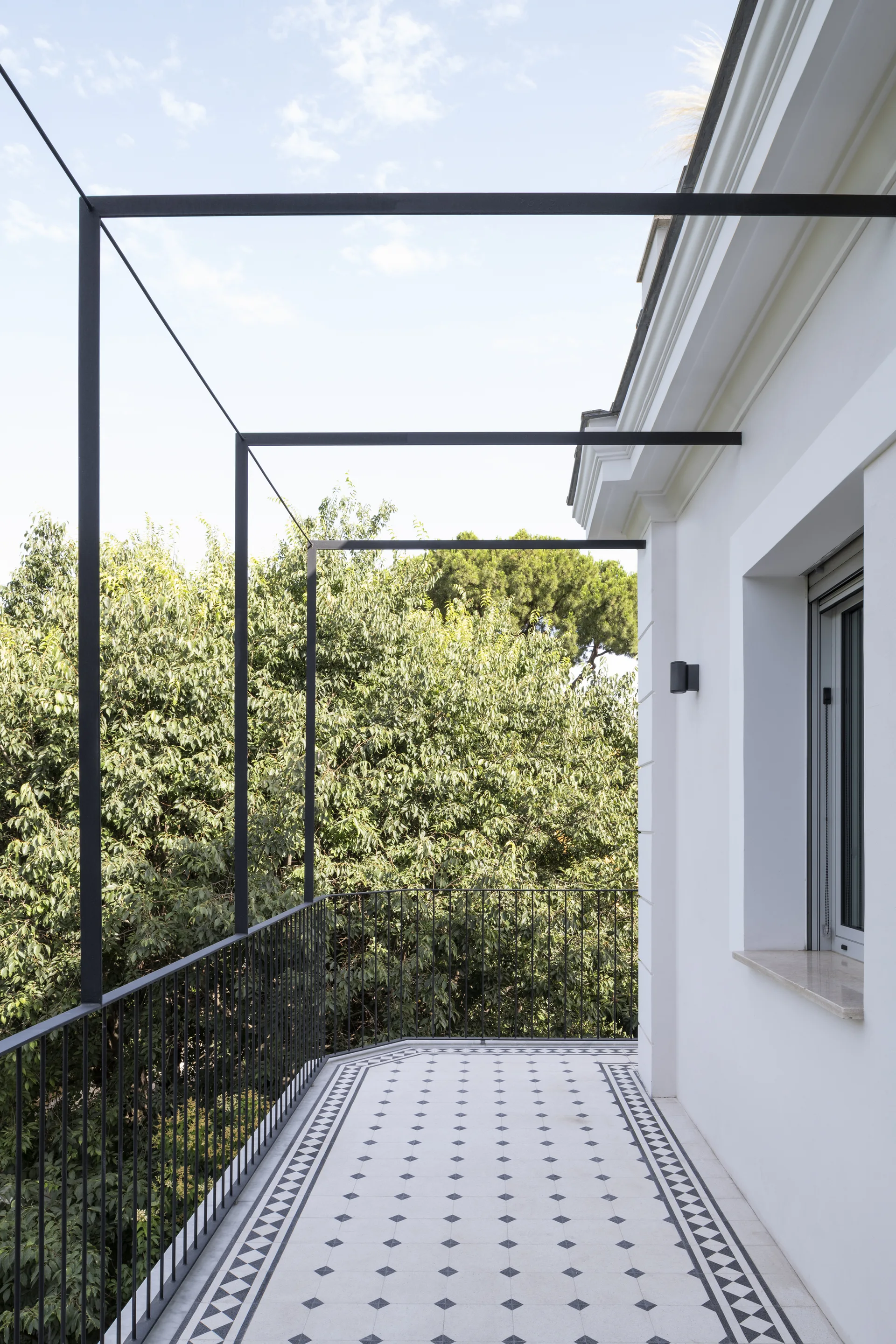 A modern balcony features geometric black and white floor tiles, a minimalist black metal pergola frame, and a dark metal railing against a white exterior wall.