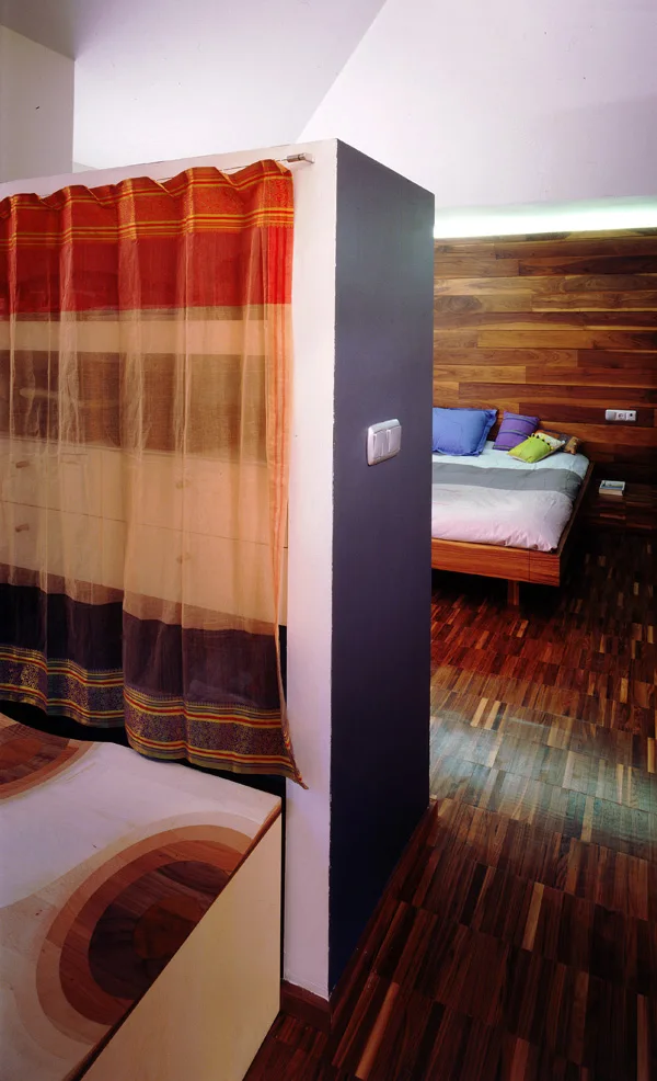 A bedroom split by a partial partition wall with a sheer orange curtain, featuring a wood paneled headboard wall and dark parquet wood flooring.