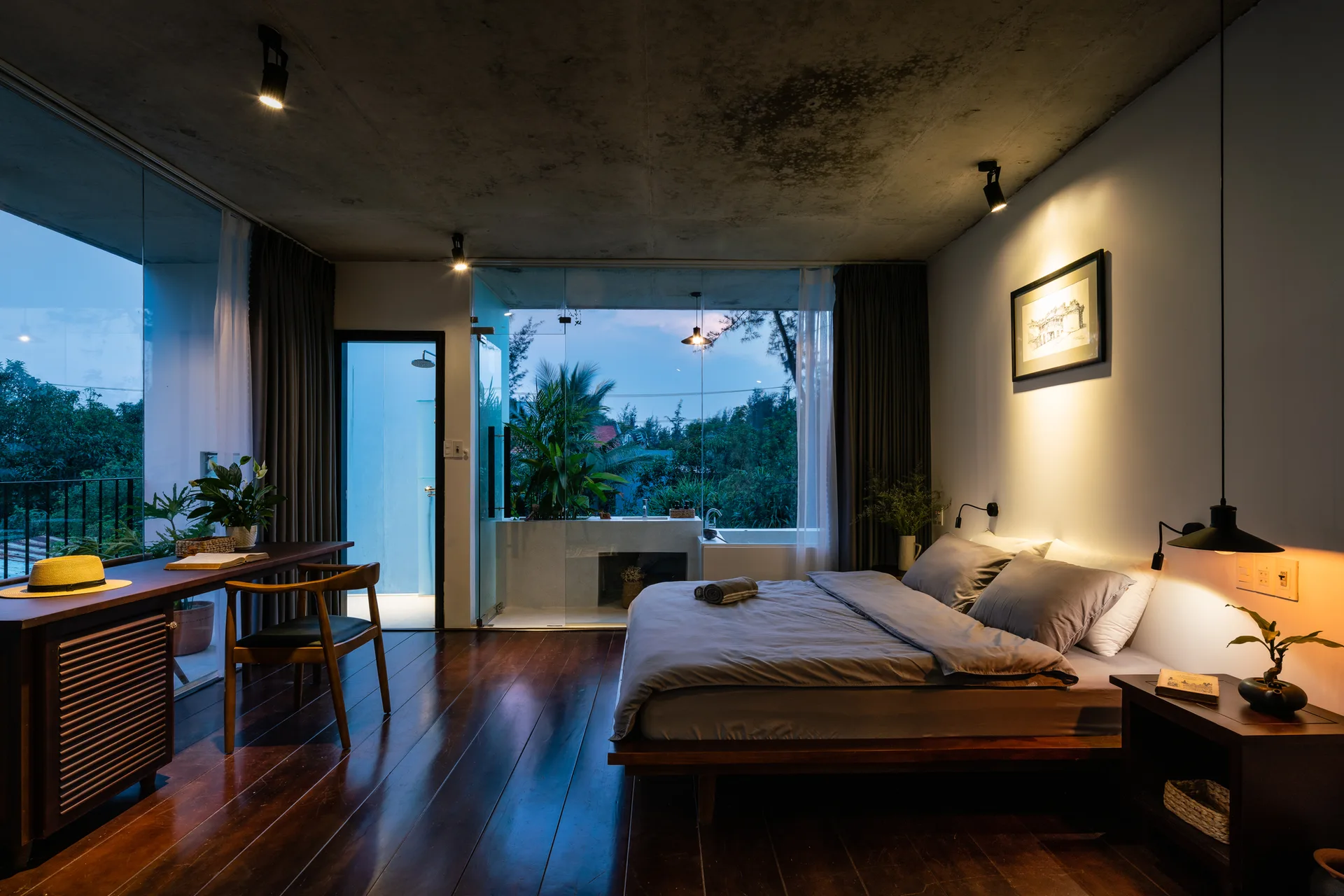 A modern bedroom features a low platform bed, a wooden desk with a chair, a raw concrete ceiling, and large glass windows overlooking greenery.