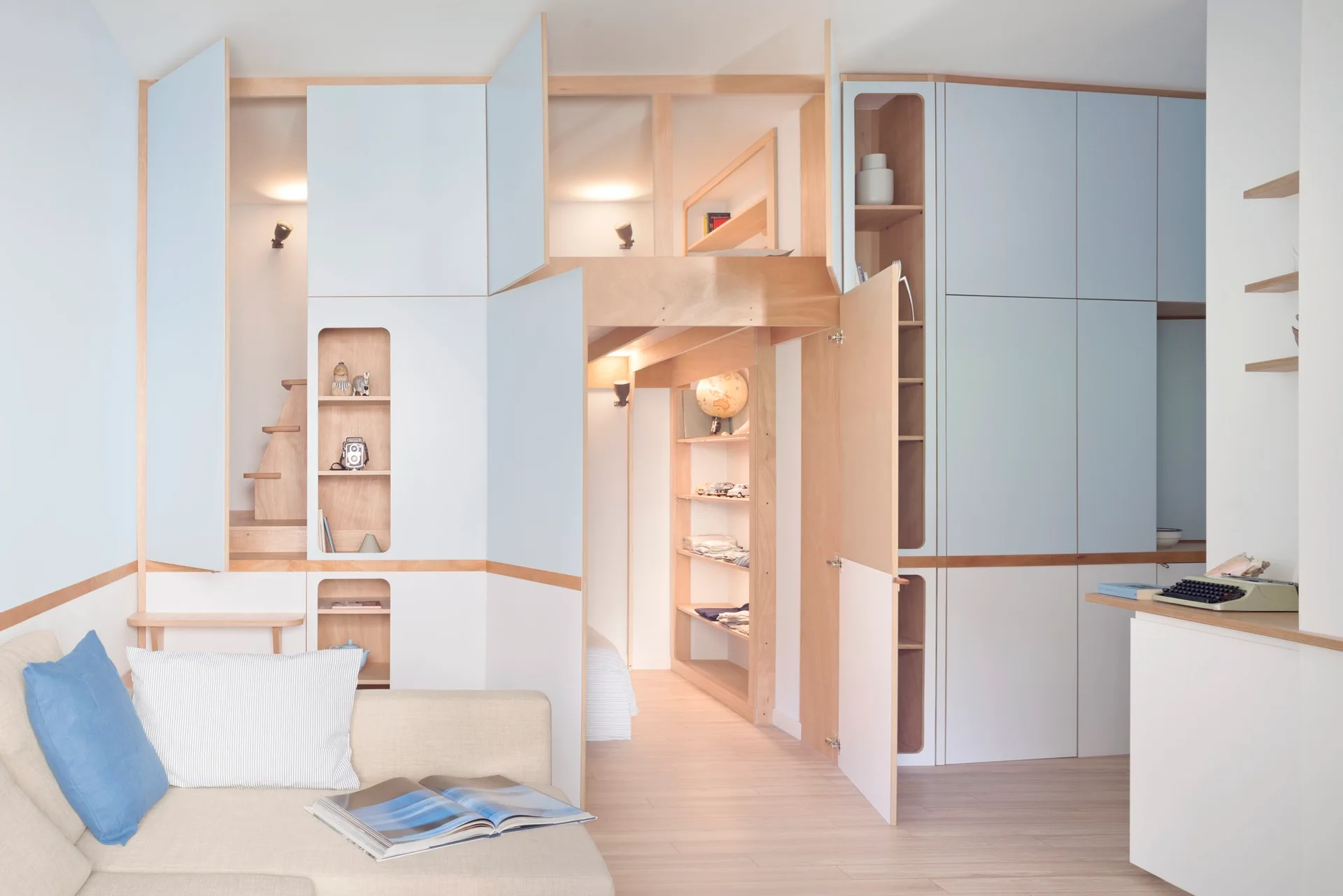 A compact studio apartment features custom light blue and wood cabinetry with a lofted bed, built-in shelving, and a neutral sofa.