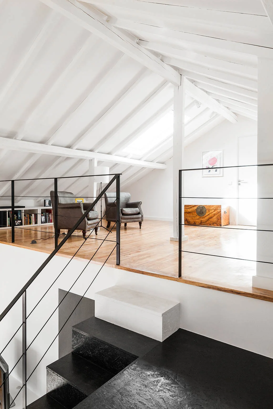 A lofted landing with a pitched white beamed ceiling, natural wood flooring, thin black metal railings, and grey leather armchairs.