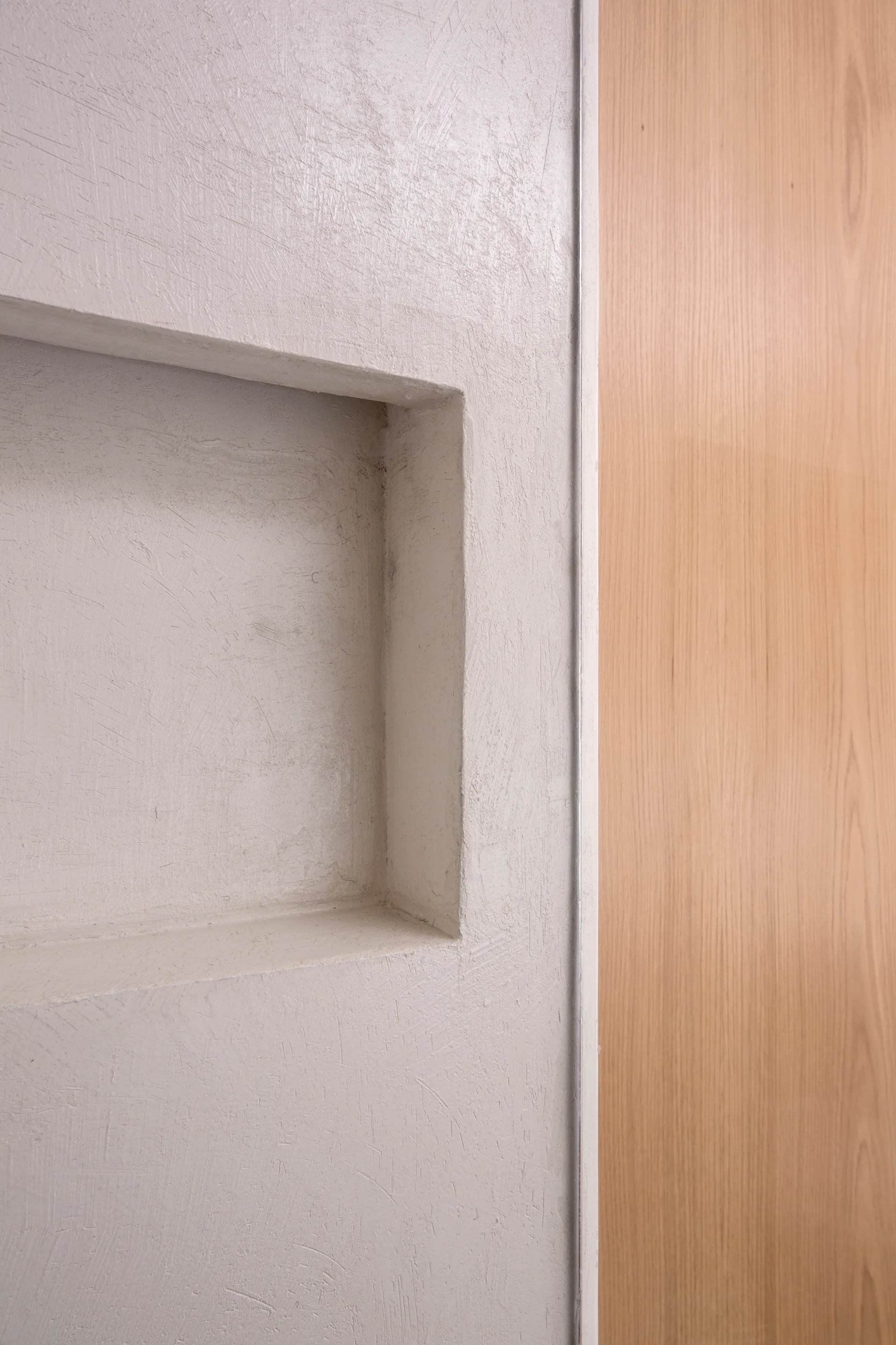 Close-up of a textured beige plaster wall featuring a recessed square niche alongside smooth vertical wood paneling separated by a thin metal trim.