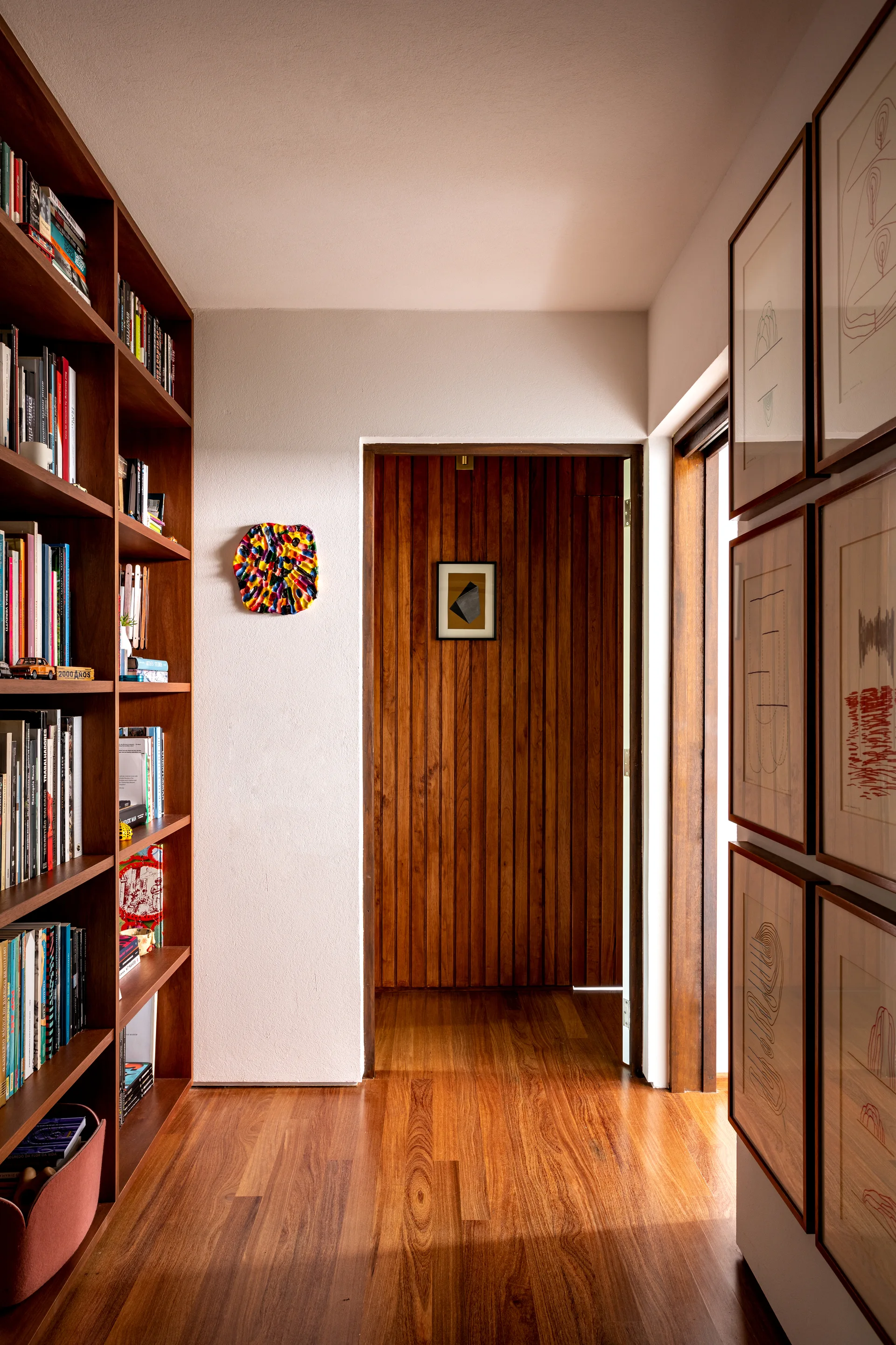 A warm wood hallway features a floor-to-ceiling built-in bookshelf, a framed gallery wall, and a vertical wood slat door at the end.