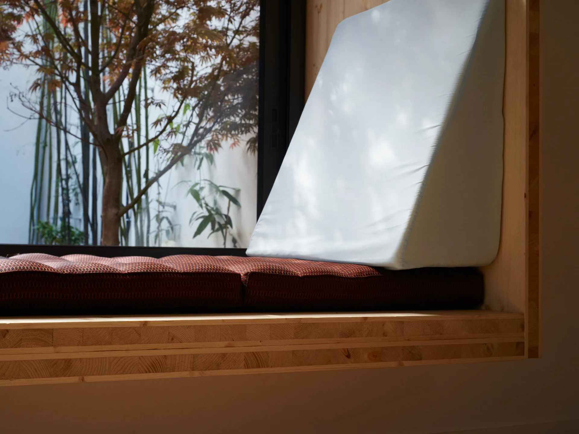 A close-up of a light wood window seat featuring a dark patterned cushion and a light blue wedge pillow, looking out onto a garden tree.