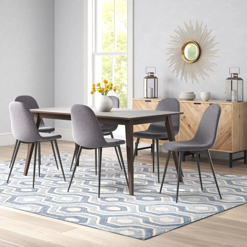 A wooden dining table and gray chairs sit on a blue geometric rug next to a light wood sideboard with a sunburst mirror.