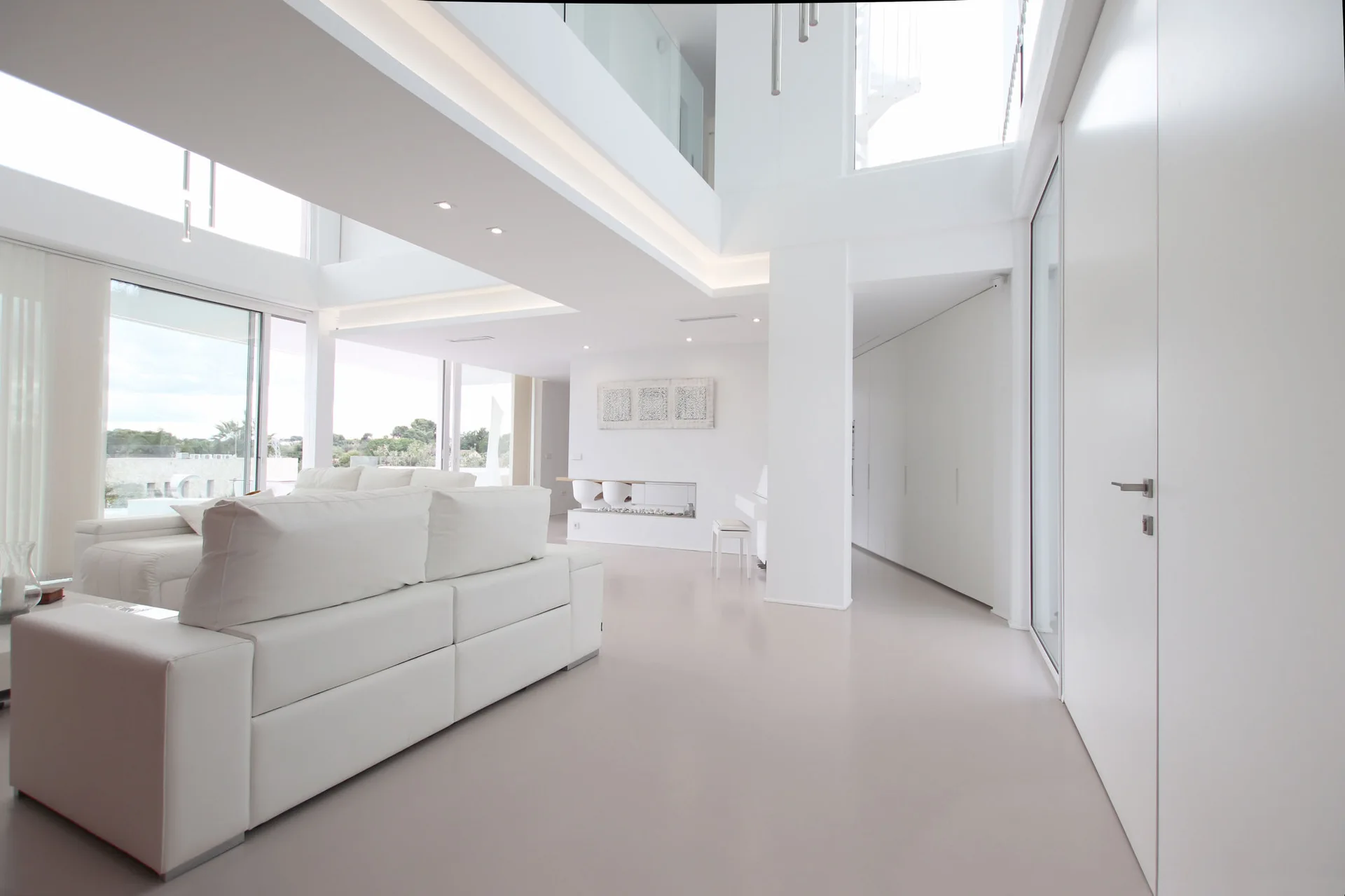 A minimalist double-height white living room with a large white sectional sofa, integrated ceiling cove lighting, and a seamless light-colored floor.
