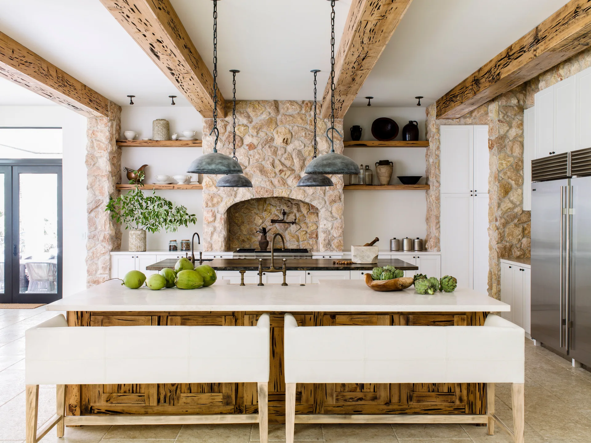A rustic kitchen features a white-topped island with wood paneling, white bar stools, exposed ceiling beams, and a large natural stone wall.