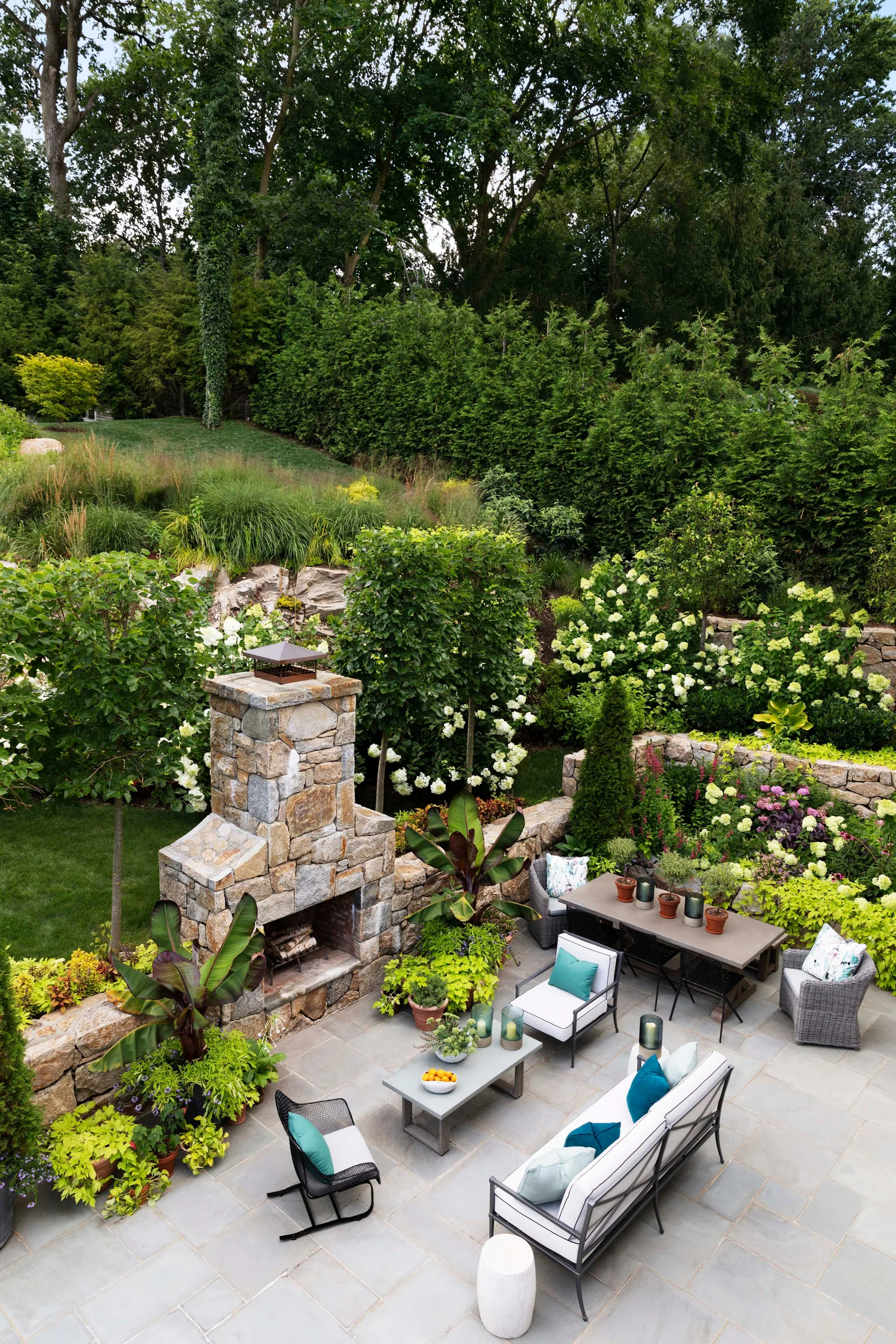 An outdoor stone patio features a large stone fireplace, grey metal and wicker furniture with teal pillows, and tiered gardens with lush green plants.