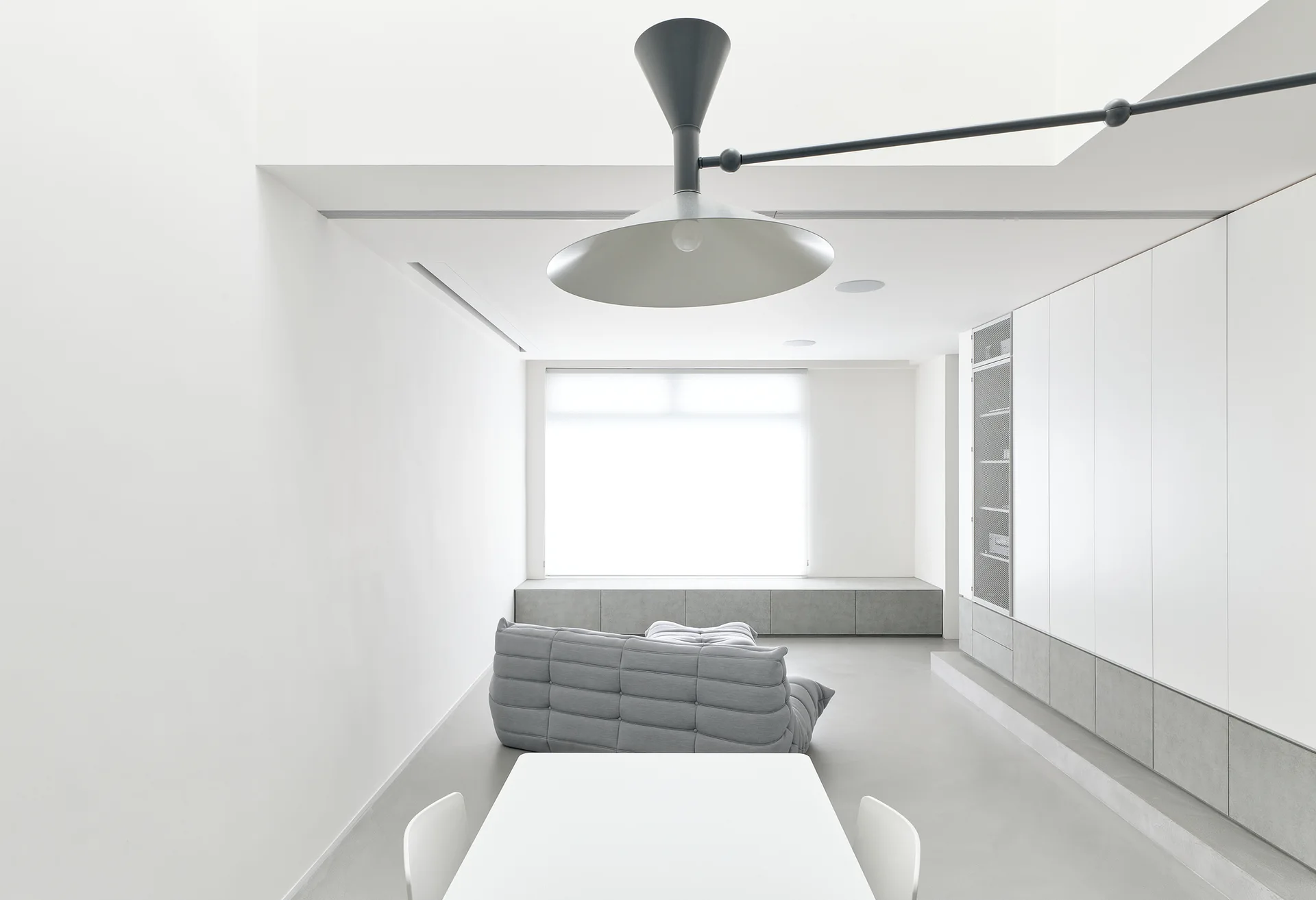 A minimalist white living room features a low grey tufted sofa, integrated wall cabinetry, a white dining table, and a large sculptural pendant light.