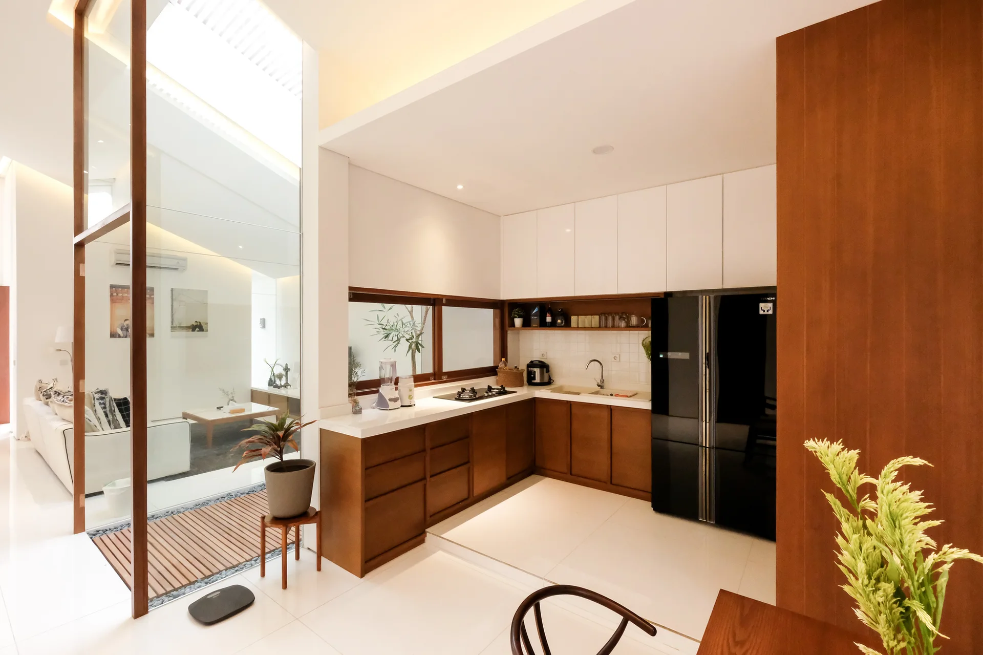 A modern kitchen with wood and white cabinetry, a black refrigerator, and a tall glass partition looking into a bright living room.