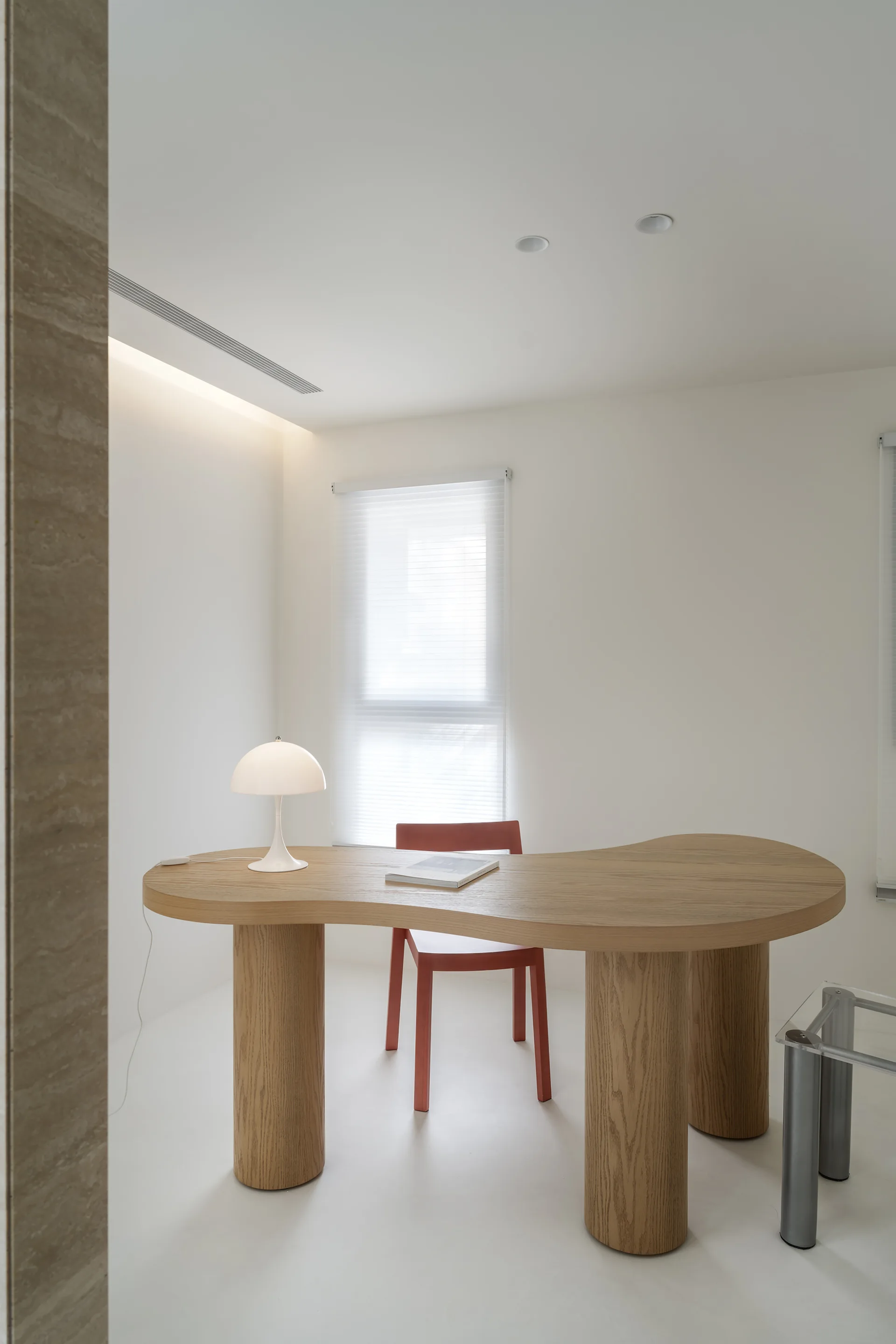 A curved wooden desk with cylindrical legs holds a white mushroom lamp next to a red chair in a minimalist white home office.