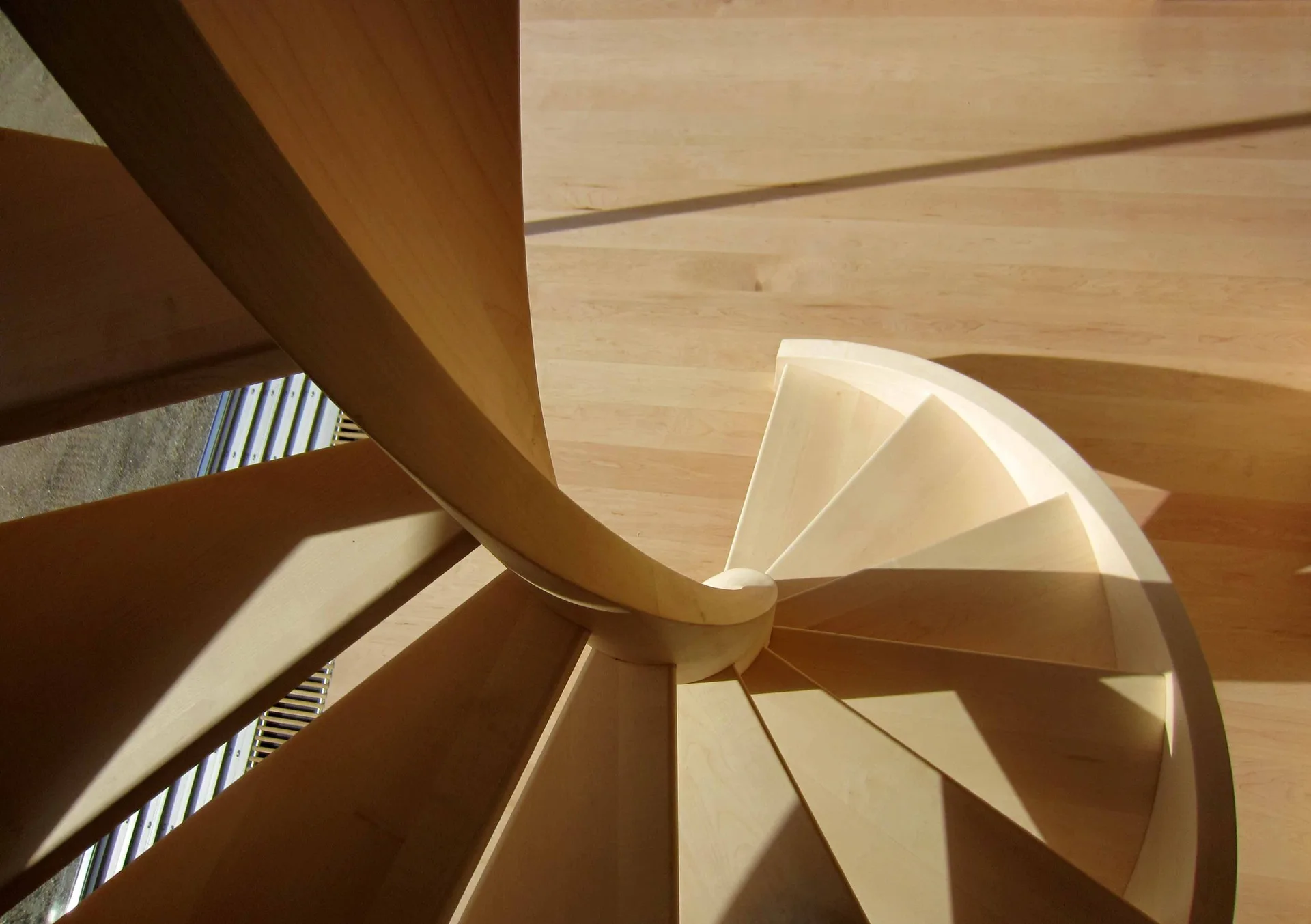 A top-down view of a light blonde wooden spiral staircase curving down to meet matching light-colored wood plank flooring.