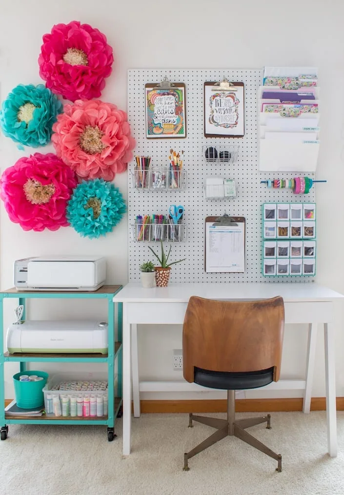 A white desk features a brown wood swivel chair, a white pegboard organizer, a teal utility cart, and large colorful paper flowers on a white wall.