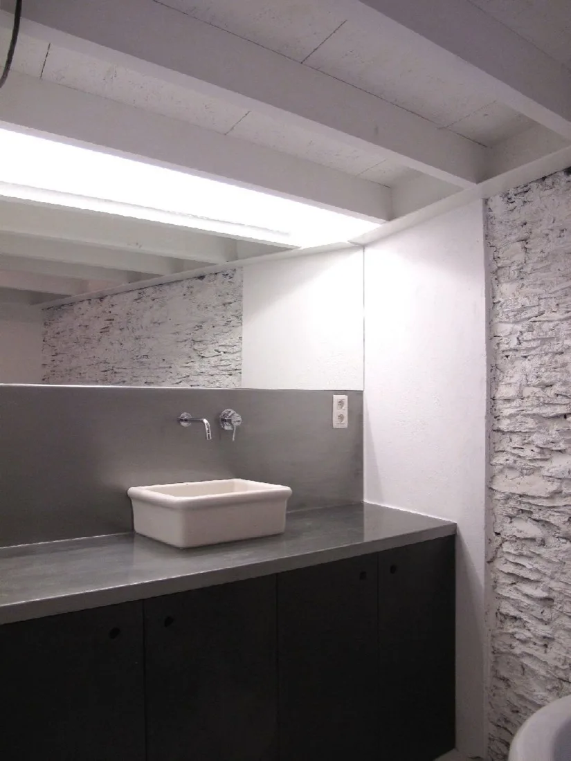 A minimalist bathroom vanity features a white vessel sink on a stainless steel countertop with a matching metal backsplash and a painted stone wall.