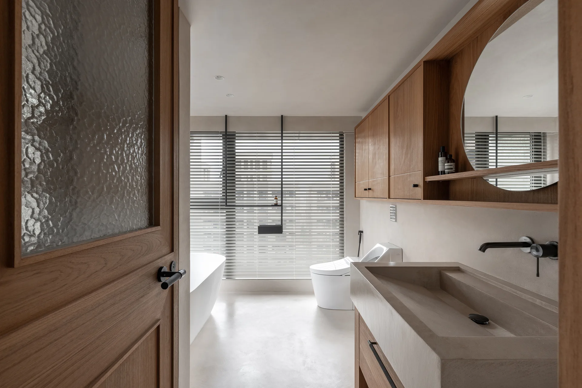 A minimalist bathroom viewed through a wood door, featuring a concrete basin sink, warm wood cabinets, a round mirror, and a freestanding bathtub.