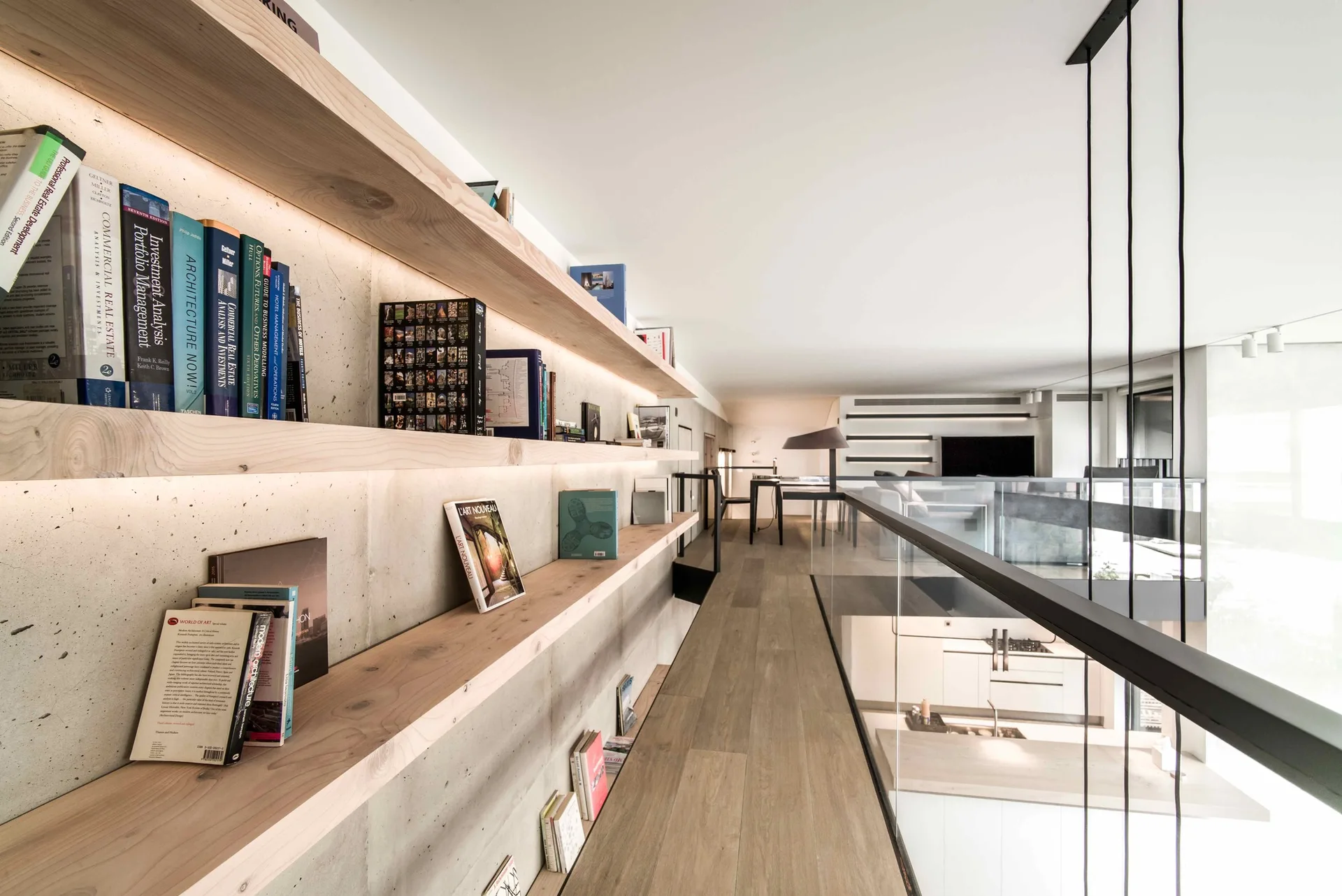 An elevated home landing with light wood floors, an exposed concrete wall with illuminated floating bookshelves, and a modern glass railing.