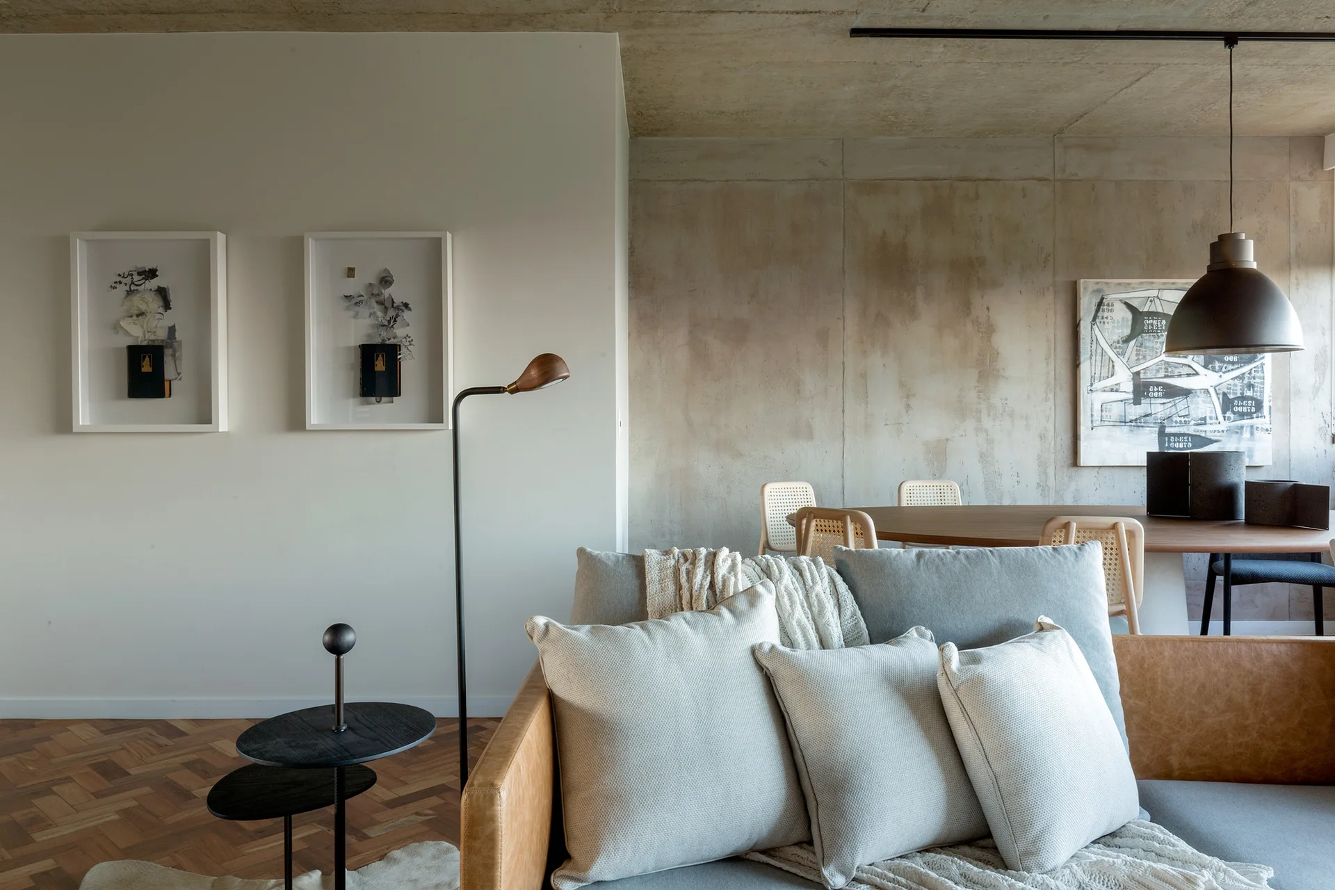 A living room featuring a tan leather sofa with gray cushions, a raw concrete wall, parquet wood flooring, and a dining table in the background.