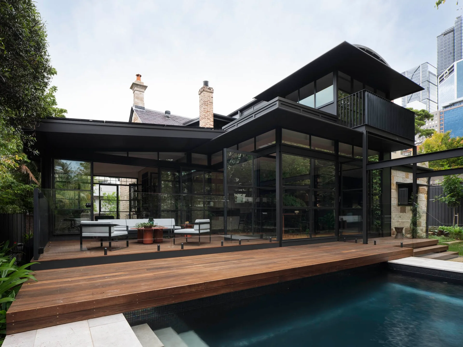 A modern patio features a wooden deck with grey lounge furniture next to a swimming pool and a black-framed glass home extension.