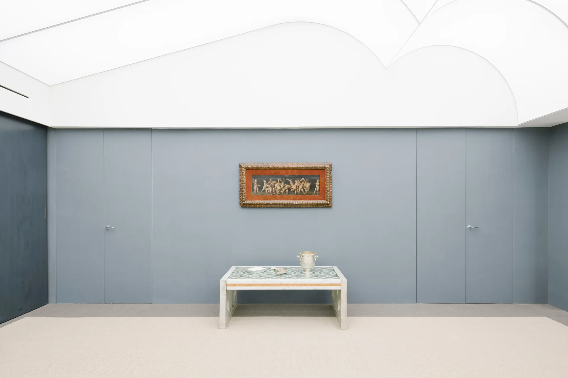 A minimalist grey-blue paneled wall features two flush concealed doors flanking a low console table and a framed painting under a scalloped white ceiling.