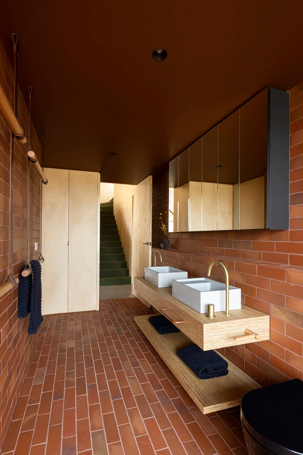 A modern bathroom featuring red brick walls and floors, a floating light wood double vanity, rectangular vessel sinks, and brass faucets.