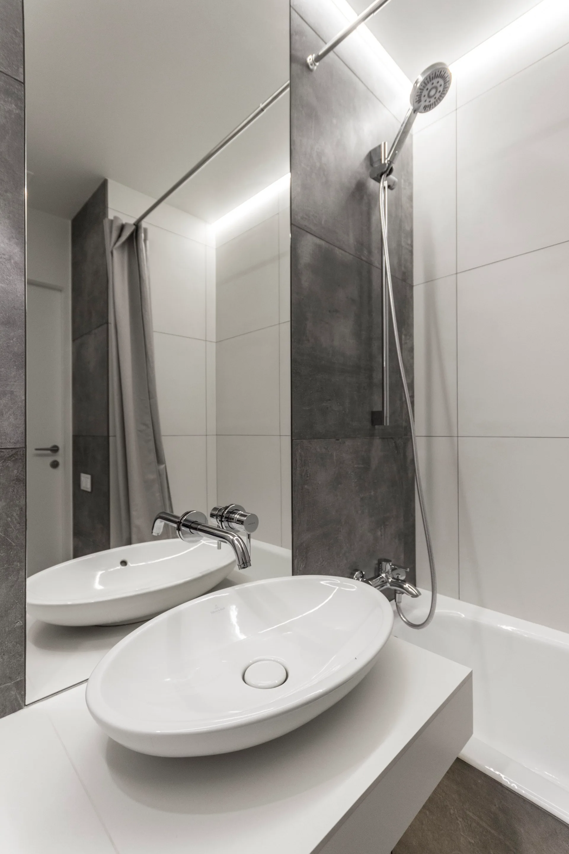 A white vessel sink sits on a floating vanity next to a bathtub, surrounded by large grey and white wall tiles and a mirrored wall.