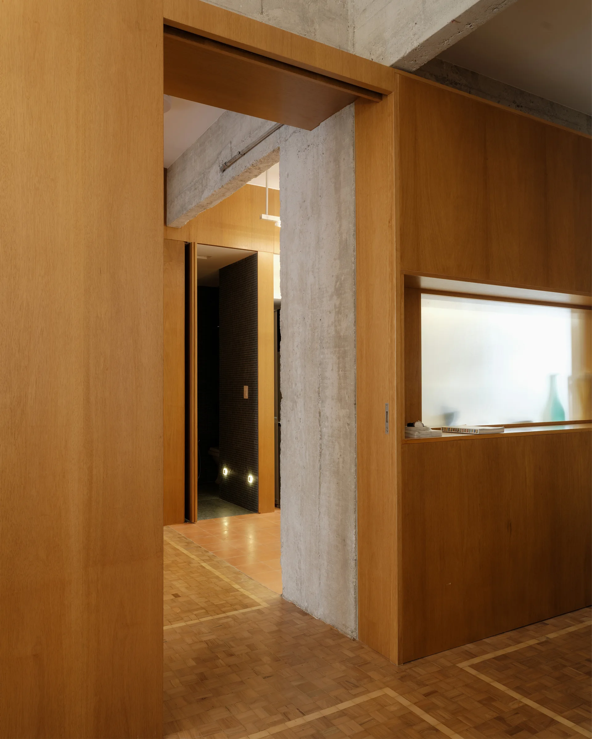A modern hallway features extensive warm wood wall paneling, parquet flooring, and a raw concrete structural column leading to another room.