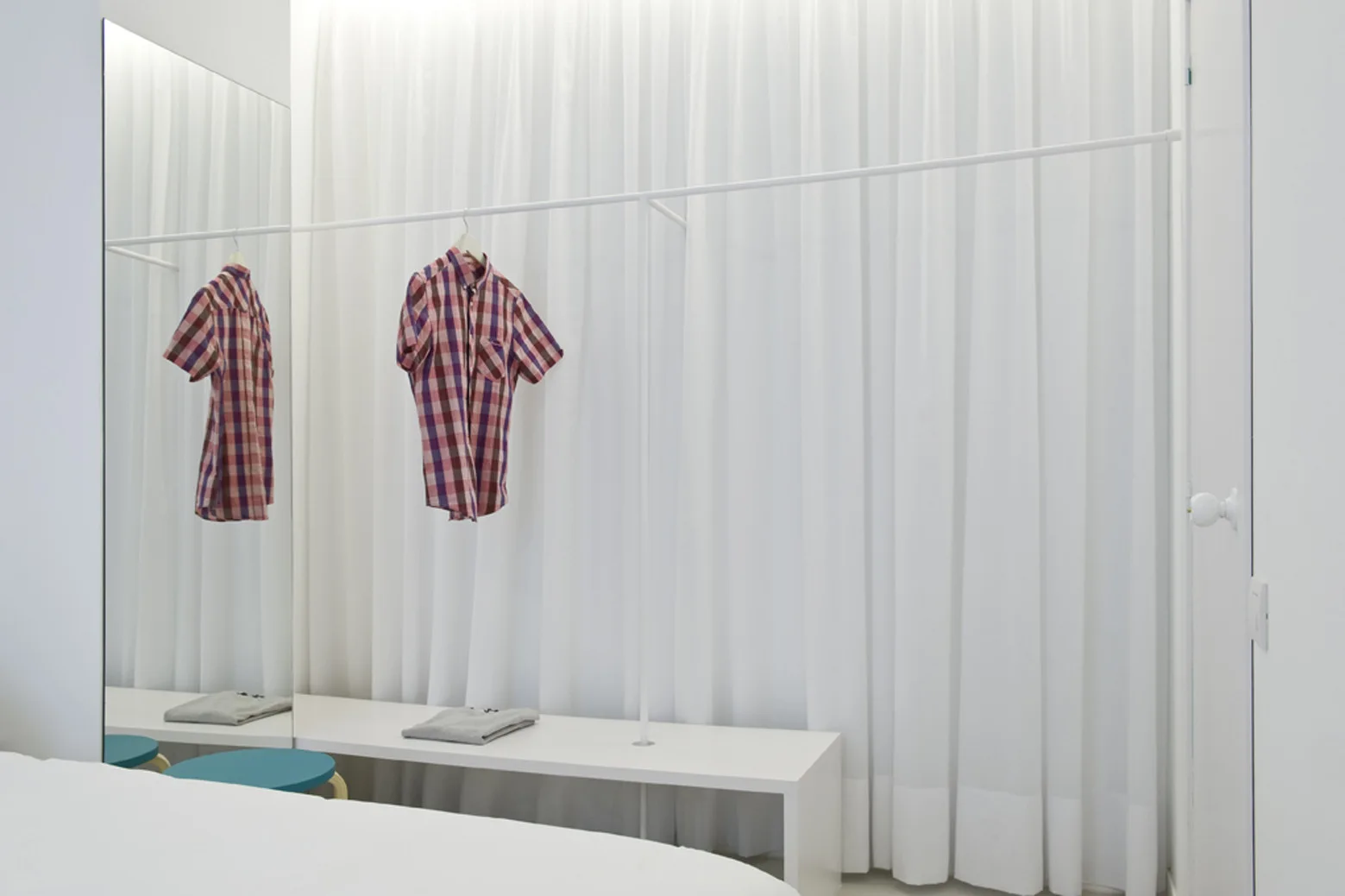 A minimalist white open closet area features a white metal clothing rack, a low bench, and a large mirror against a white curtain backdrop.