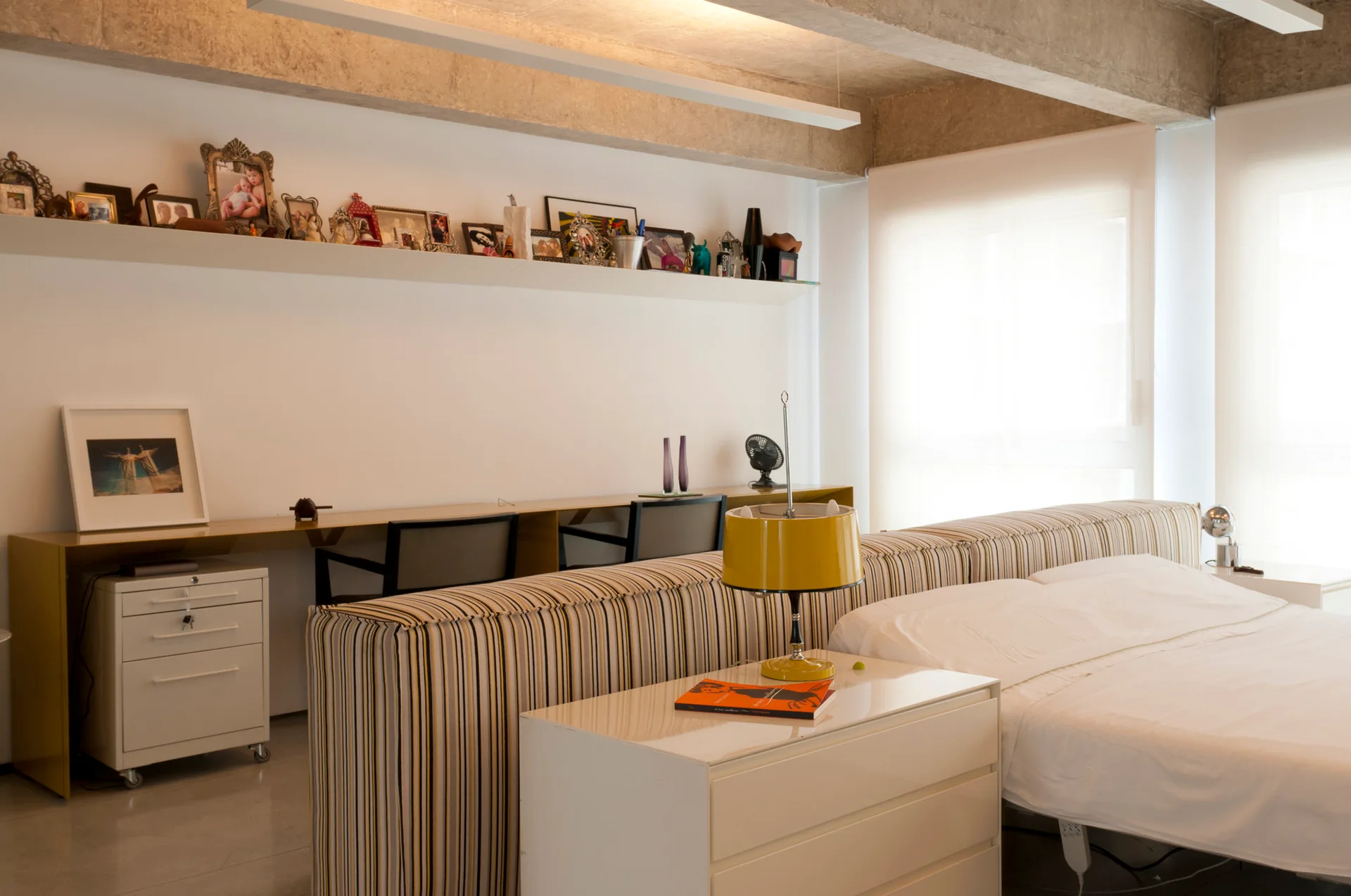 A contemporary bedroom featuring a striped upholstered room divider behind the bed and a long built-in study desk with chairs.