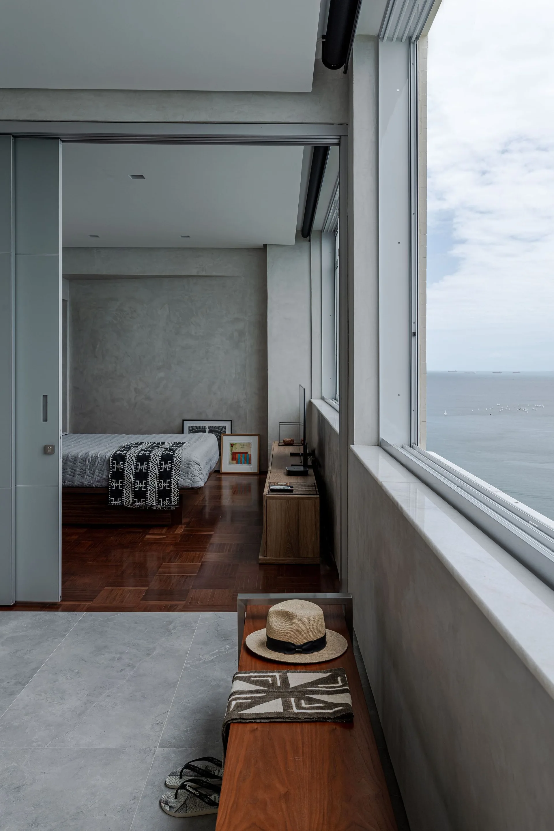 A minimalist bedroom featuring textured concrete walls, dark parquet wood floors, and a large window overlooking the ocean.