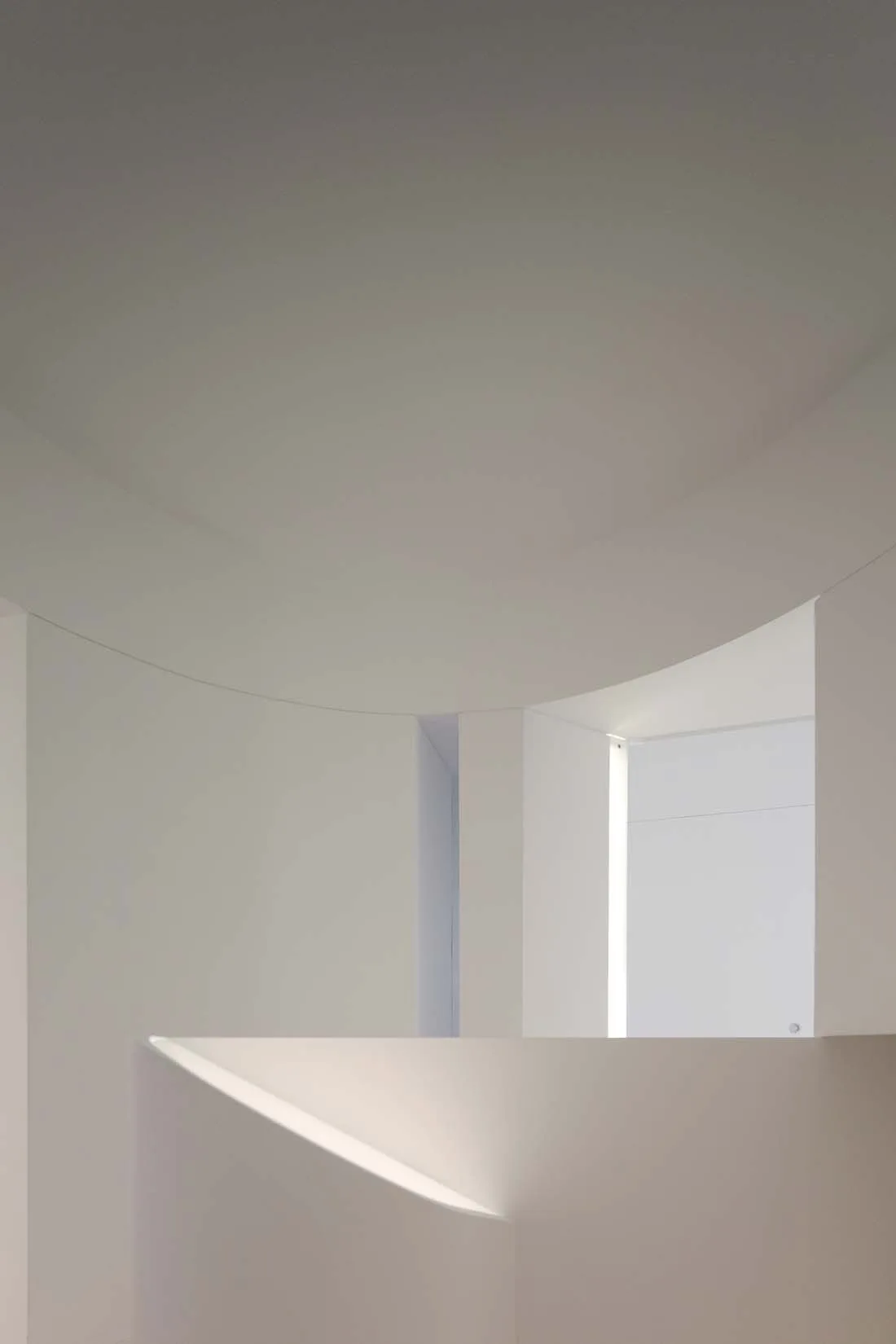 A minimalist white architectural space featuring a smooth curved plaster wall and a recessed vertical light strip.