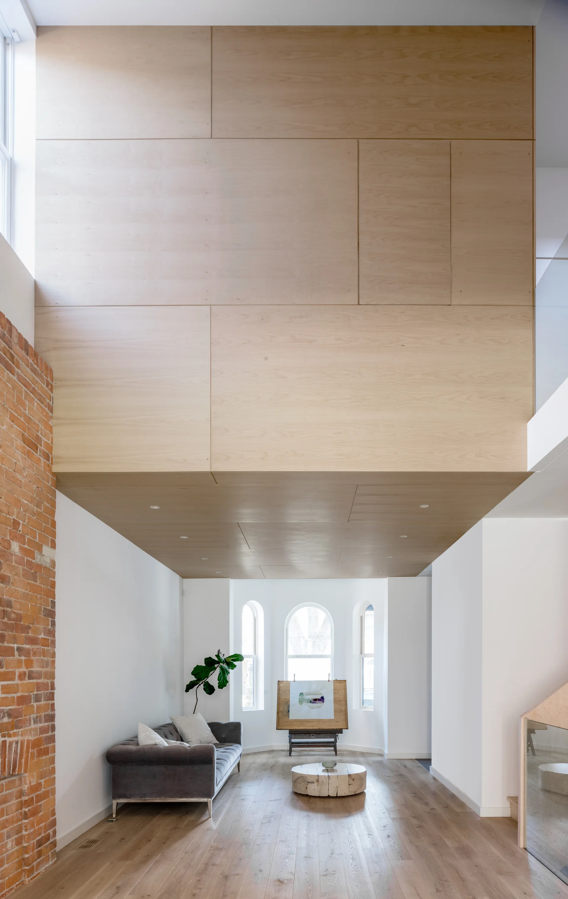 A modern living room featuring a massive light wood paneled wall above a grey sofa, flanked by an exposed brick wall and arched windows.