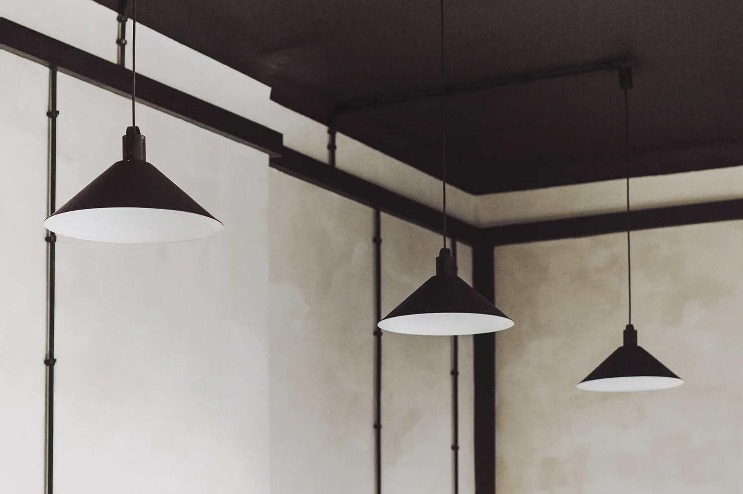 Three matte black cone-shaped pendant lights hang from a black ceiling against off-white textured plaster walls accented with vertical conduit piping.