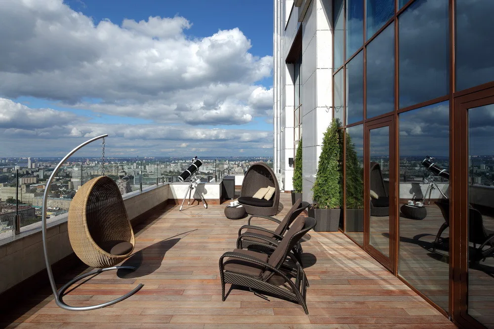 A wood-planked high-rise terrace features a wicker hanging egg chair, lounge chairs, tall potted evergreens, and panoramic views of a city skyline.
