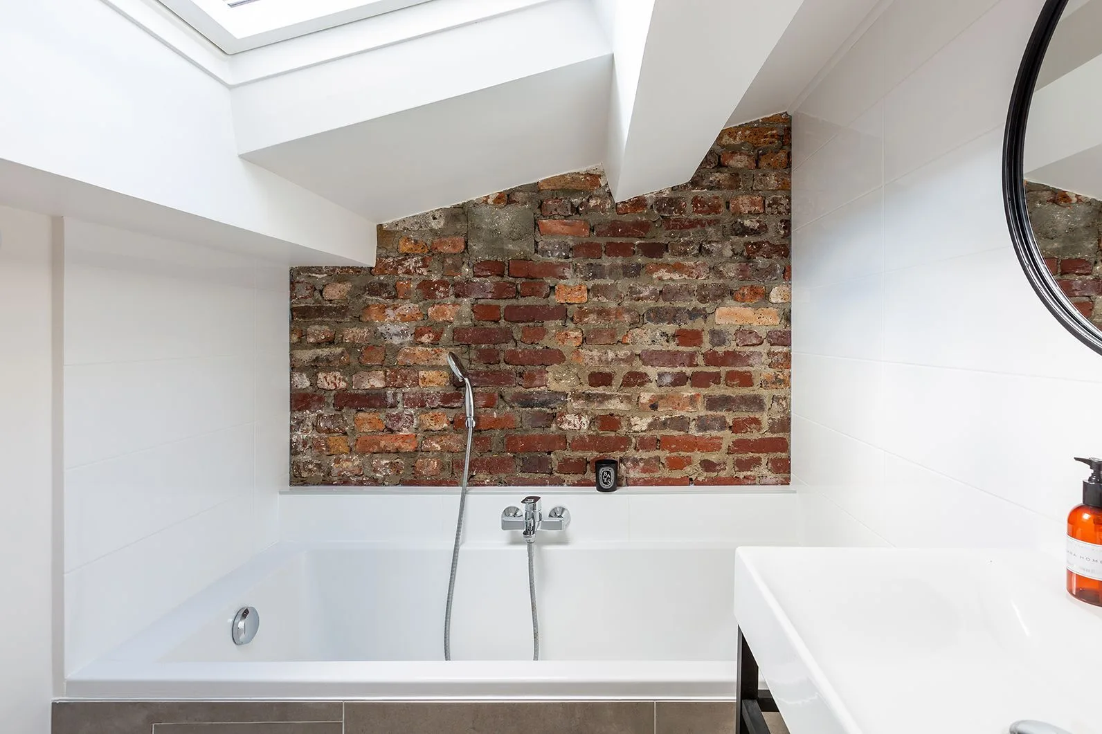 A white built-in bathtub sits under an angled skylight, positioned against an exposed red brick accent wall in a modern attic bathroom.