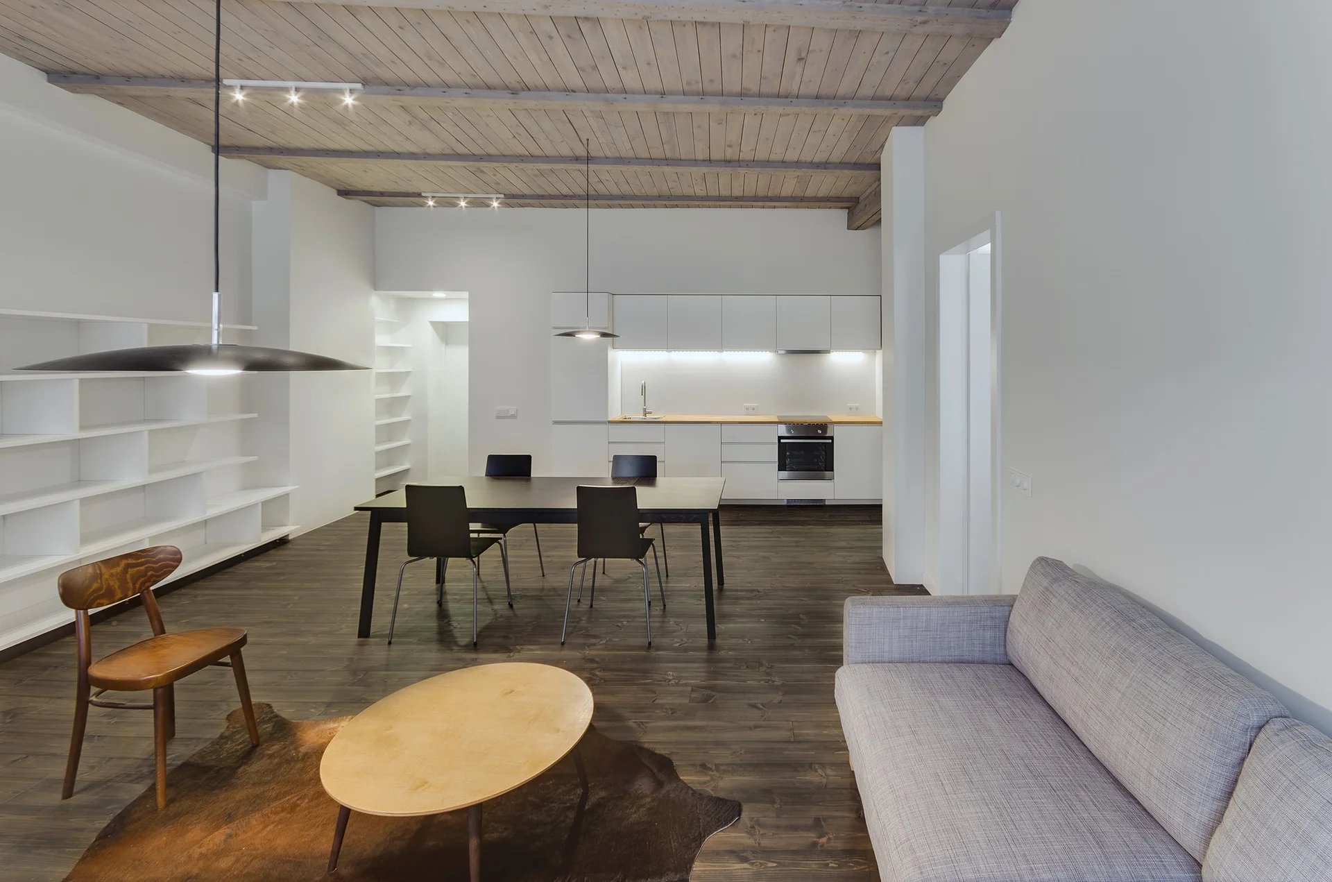 An open plan living space with a grey sofa, dark dining set, white minimalist kitchen, and a light wood plank ceiling.