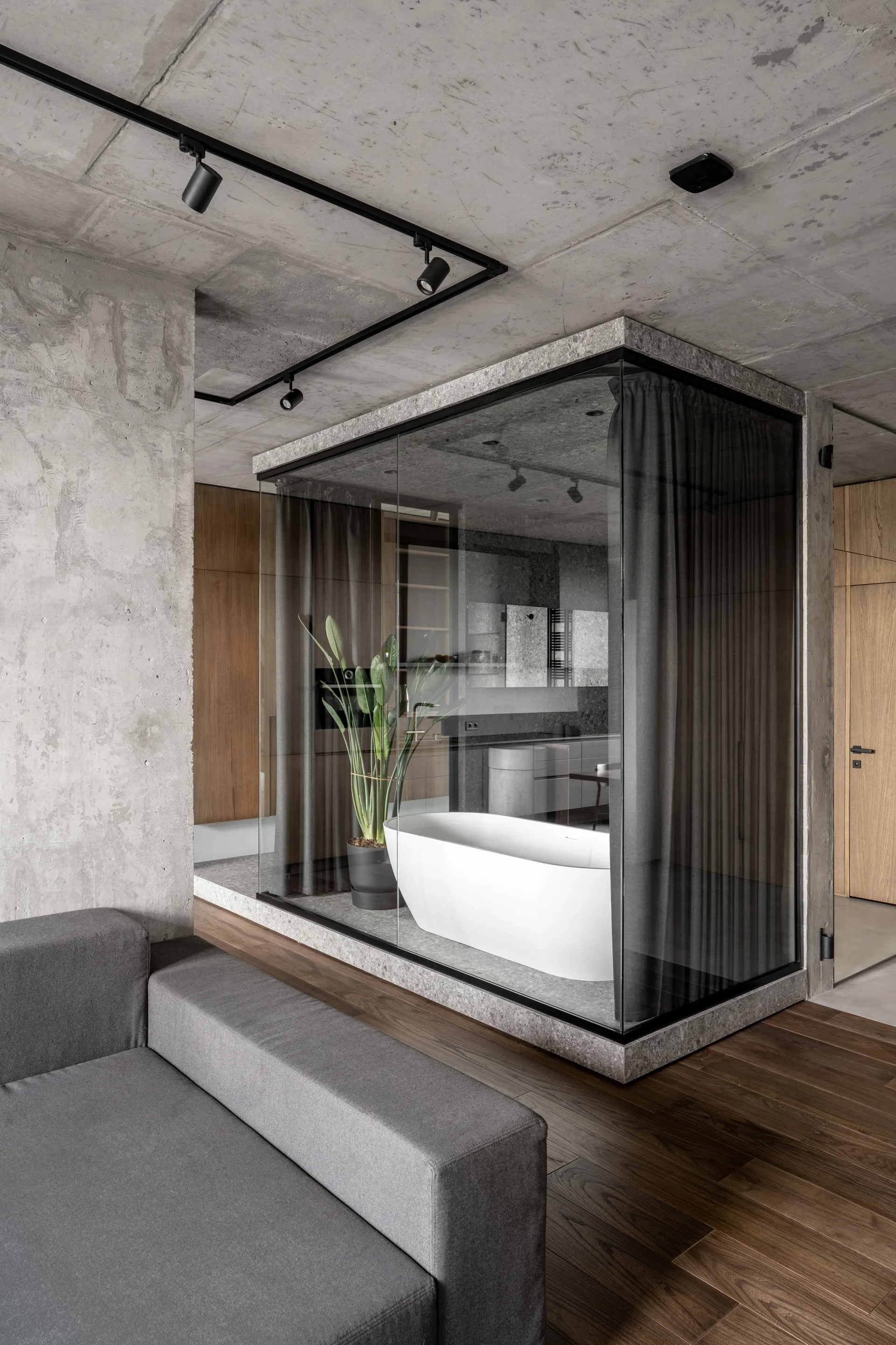 A white freestanding bathtub sits inside a glass-walled enclosure with grey curtains, surrounded by exposed concrete walls and wood flooring.