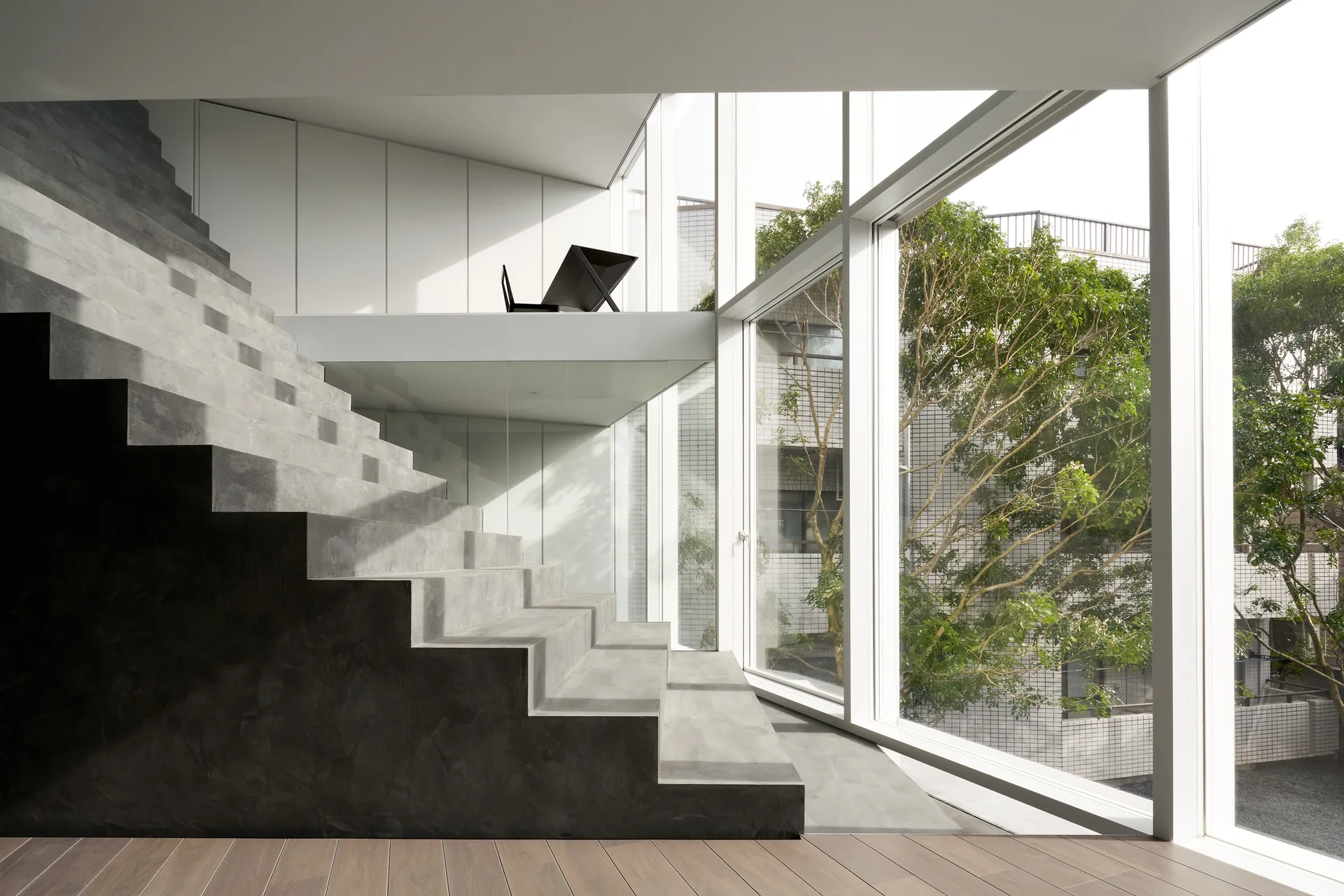 A gray concrete staircase ascends next to a tall glass window wall overlooking trees, with a white paneled wall on the mezzanine.