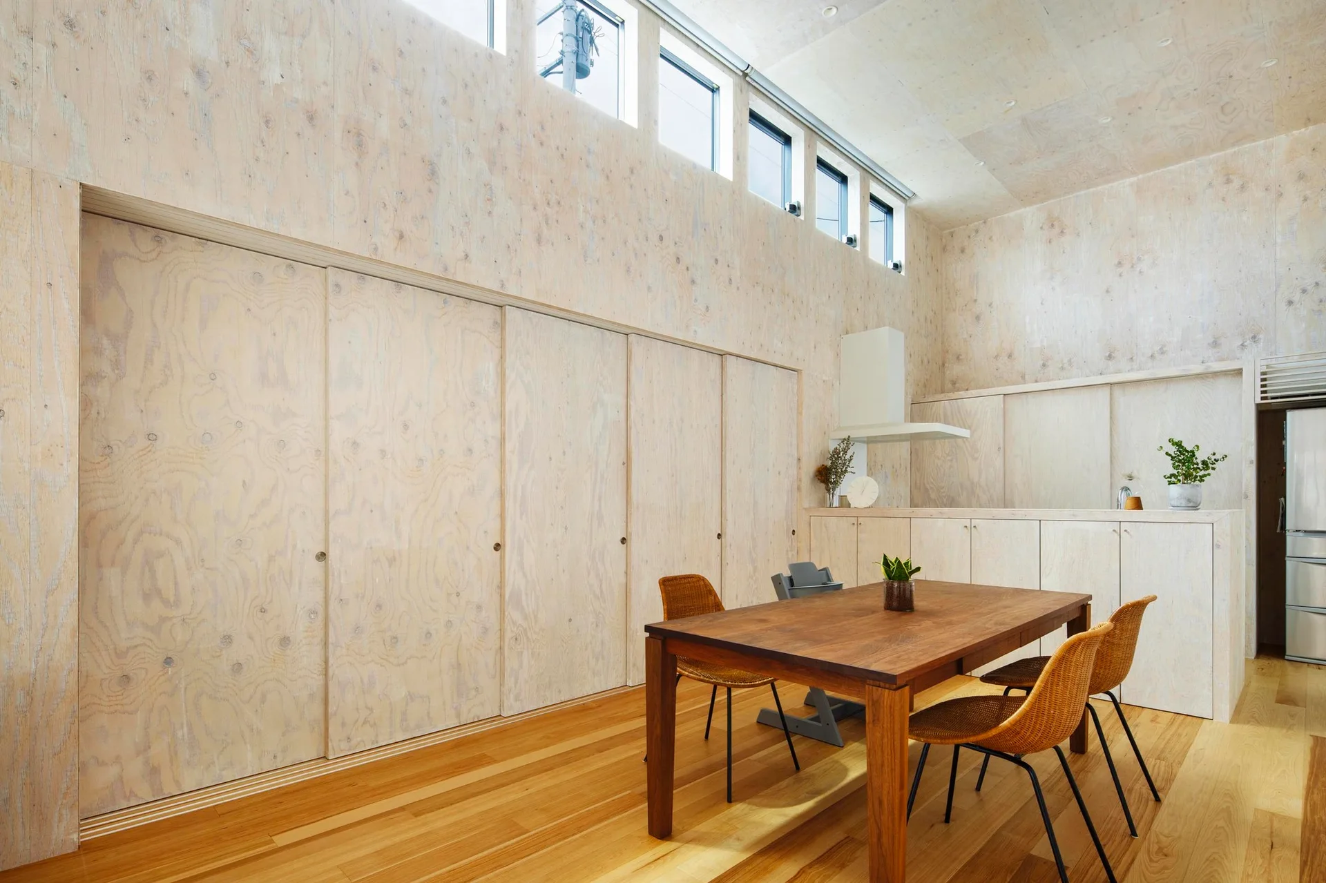 A dining room features plywood-paneled walls and ceiling, a wooden dining table with woven chairs, and a high row of clerestory windows.