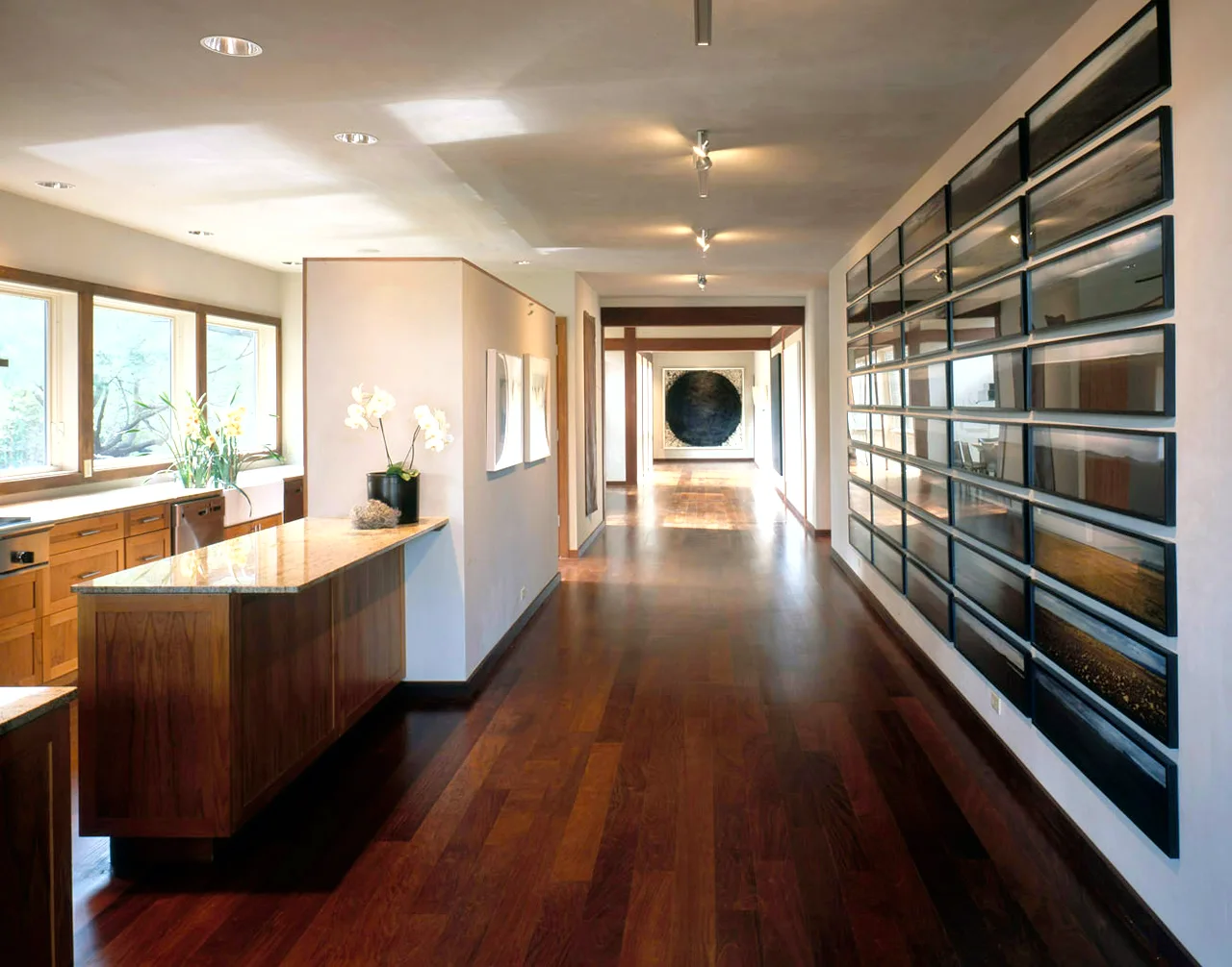 A long residential hallway featuring a extensive gallery wall of framed art, dark hardwood flooring, and a kitchen island with stone countertops.