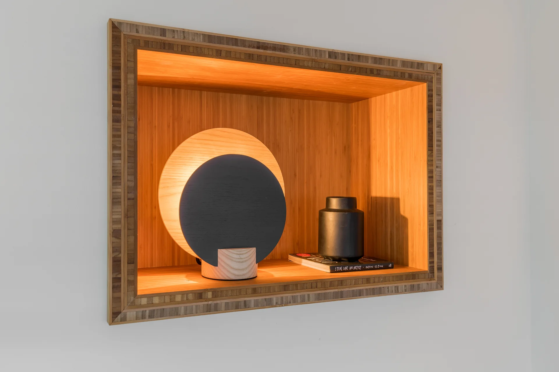 A rectangular recessed wood niche in a white wall contains a glowing circular table lamp, a book, and a matte black vase.