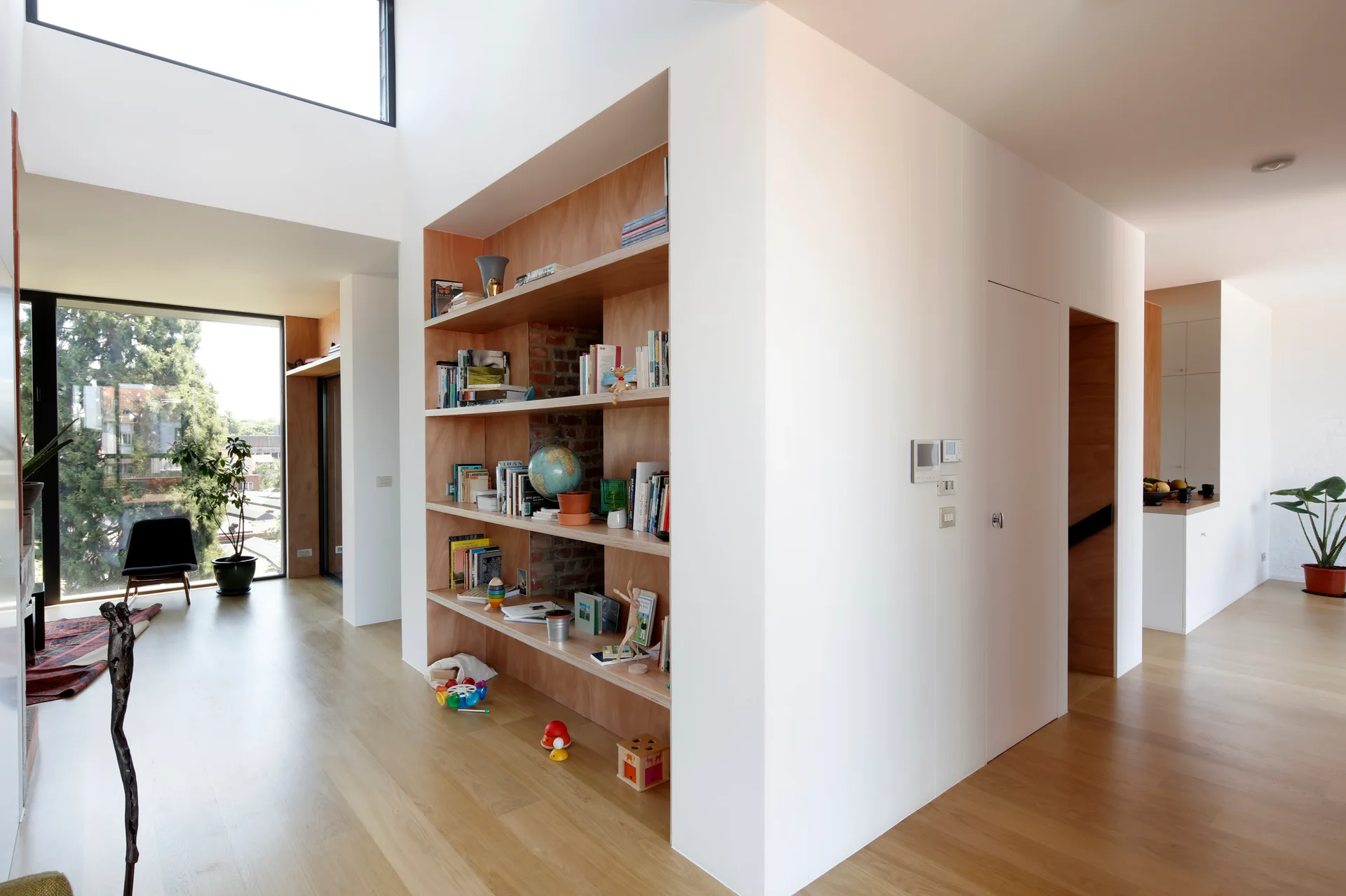 A modern home interior featuring a recessed light wood bookshelf with an exposed brick back, next to a hallway and a bright living area.