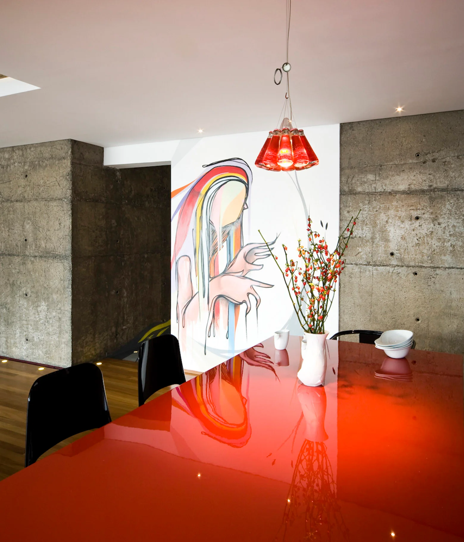 A modern dining room features a high-gloss red table, black chairs, a red bottle pendant light, and a large wall mural beside raw concrete walls.