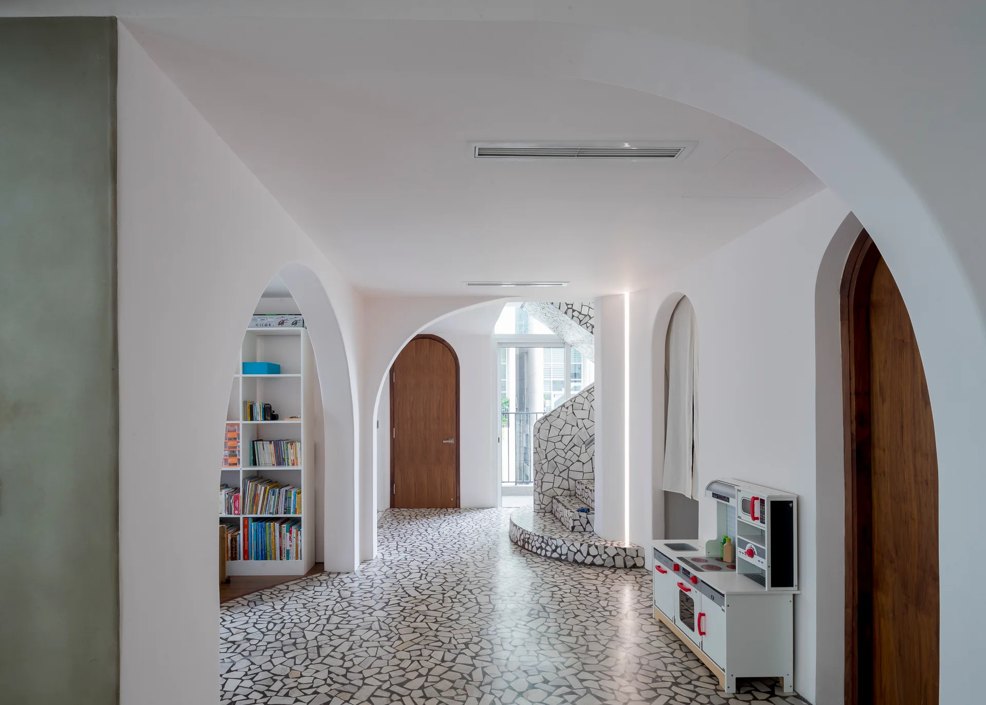 A bright minimalist hallway with white plaster archways, a curved staircase, and an irregular stone mosaic floor.