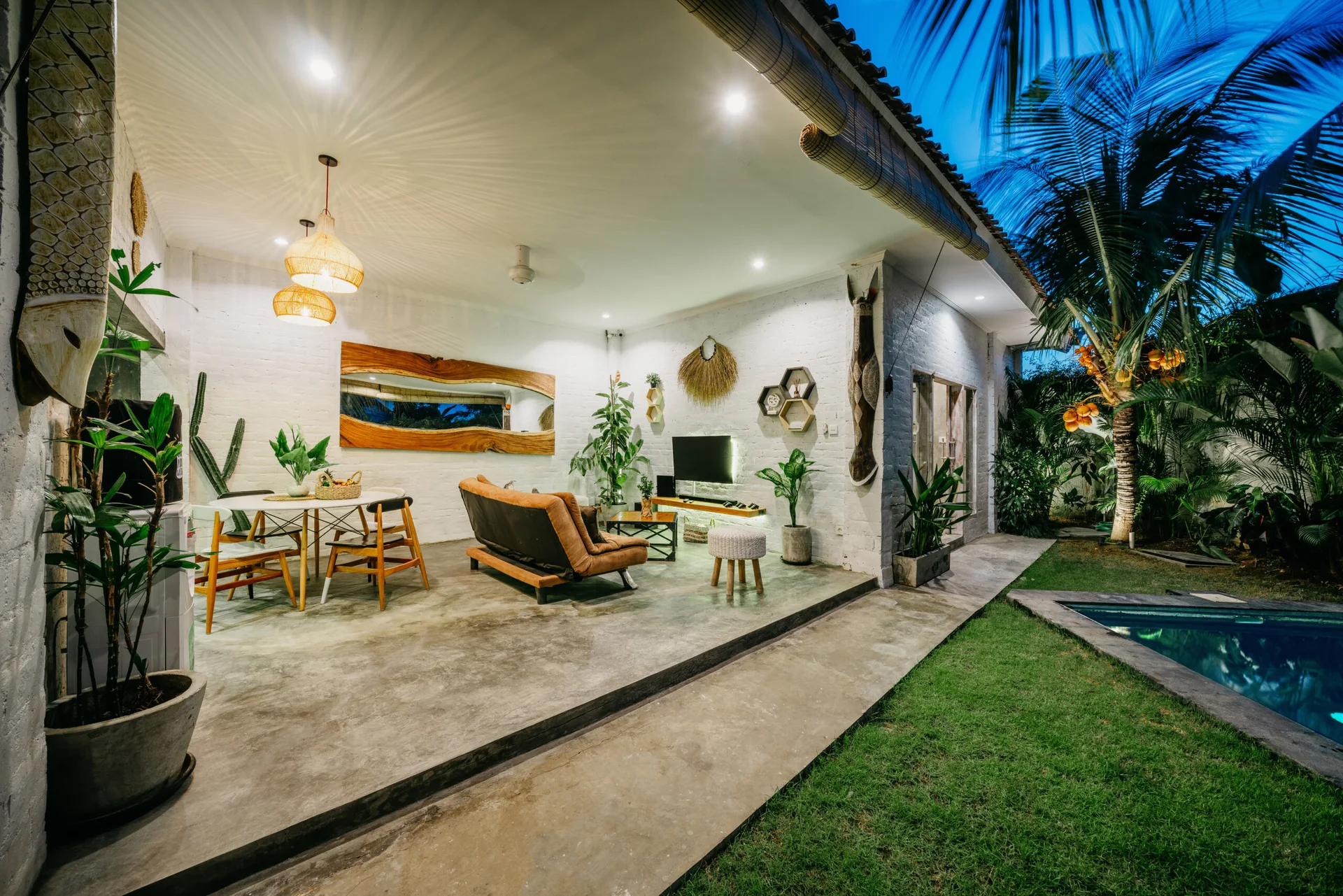A covered open-air patio with white painted brick walls, a leather sofa, and a dining table overlooks a grassy lawn and swimming pool.