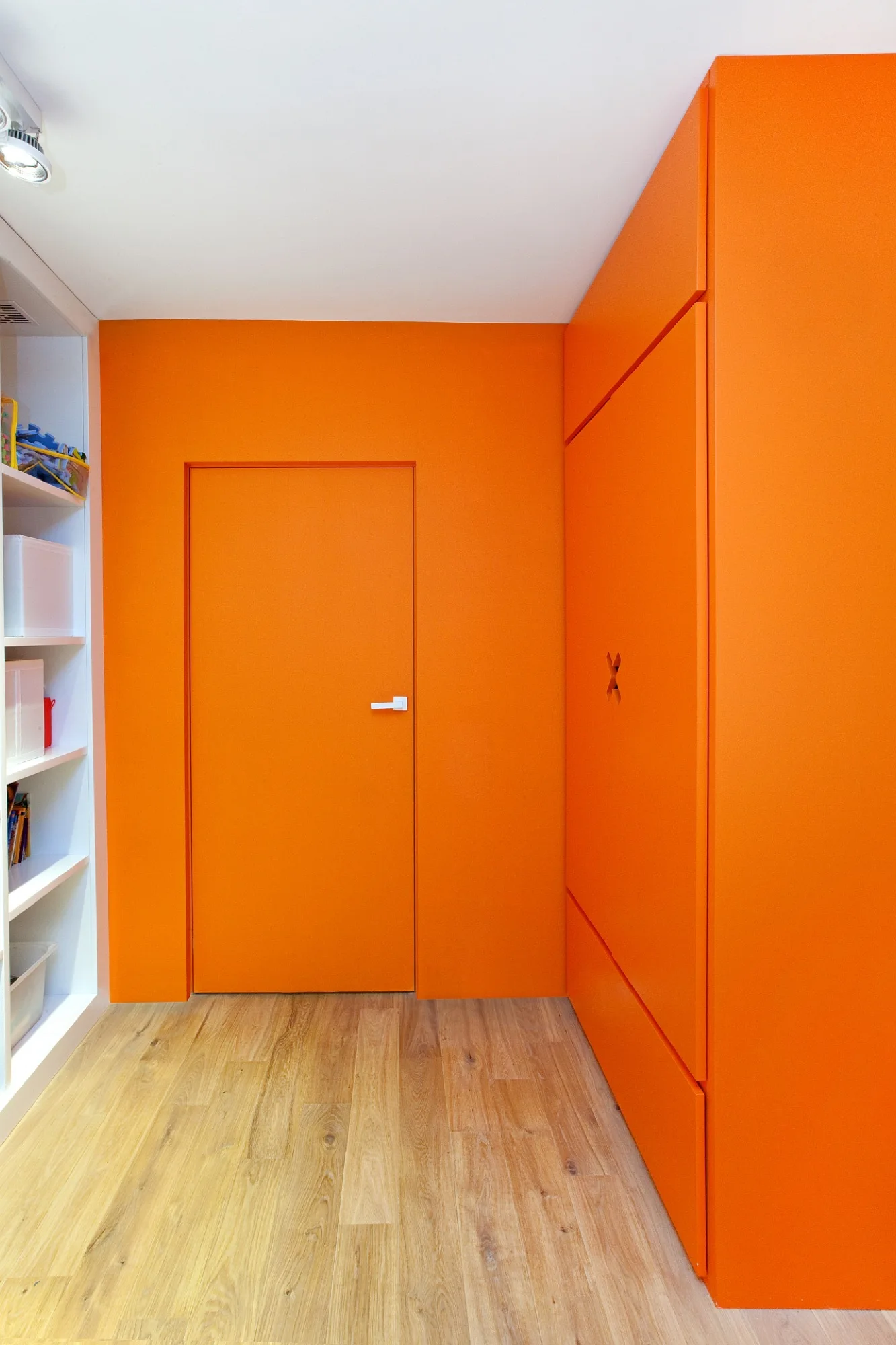 A room corner with vibrant matte orange walls, a matching integrated flush door, and orange cabinetry next to white built-in shelving.