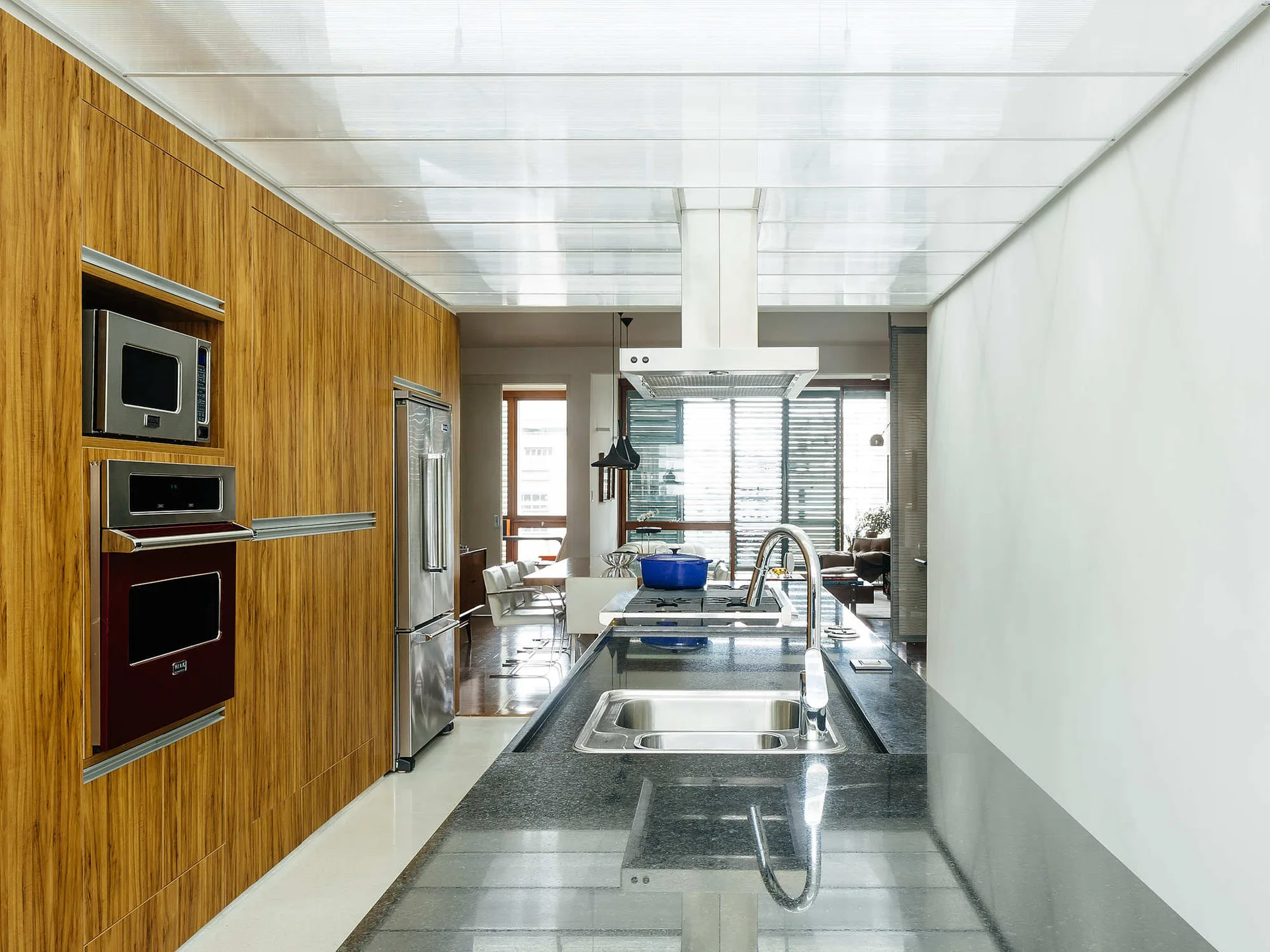 A modern kitchen featuring a long black stone island, a floor-to-ceiling wood cabinet wall, and a translucent grid ceiling.