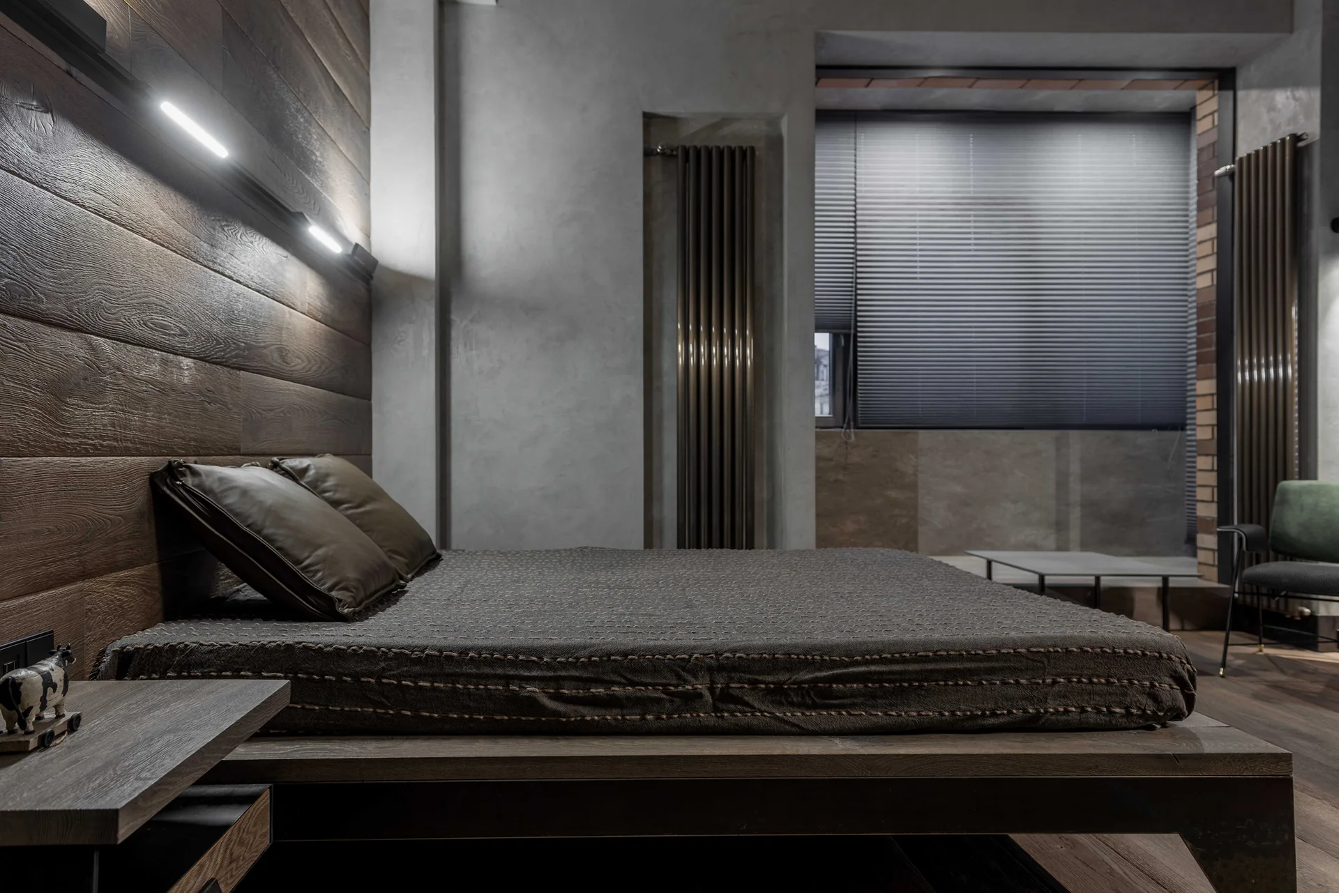 A moody bedroom featuring a dark wood platform bed against a wood-paneled wall with integrated linear lighting and raw concrete walls.